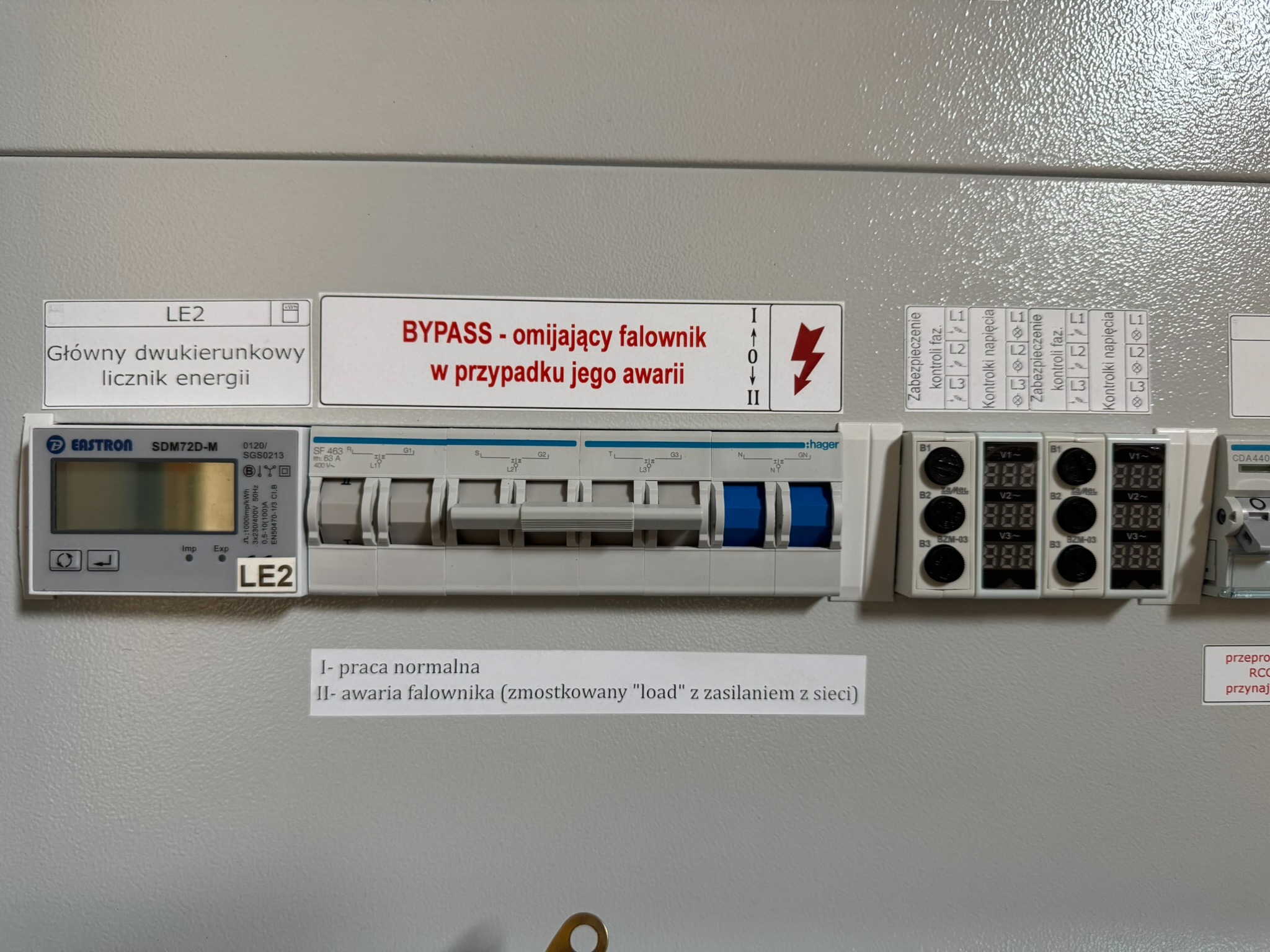 Szara szafa elektryczna z zamontowanym dwukierunkowym licznikiem energii, wyłącznikami nadprądowymi Hager z niebieskimi dźwigniami oraz modułami kontroli napięcia, oznaczona naklejkami...