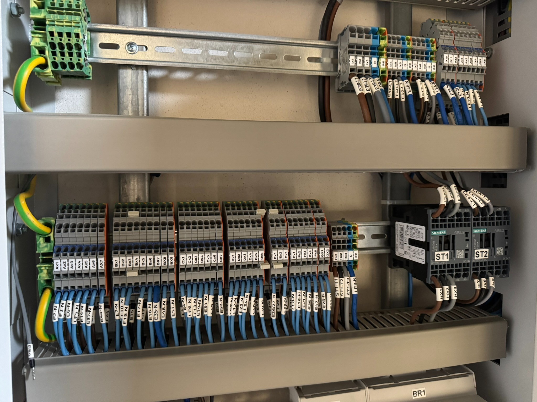 Rozdzielnica elektryczna z uporządkowanymi przewodami oznaczonymi etykietami, listwy zaciskowe Wago, styczniki Siemens Sirius i przewody ochronne żółto-zielone, zamontowane na szynach DIN w szarej...