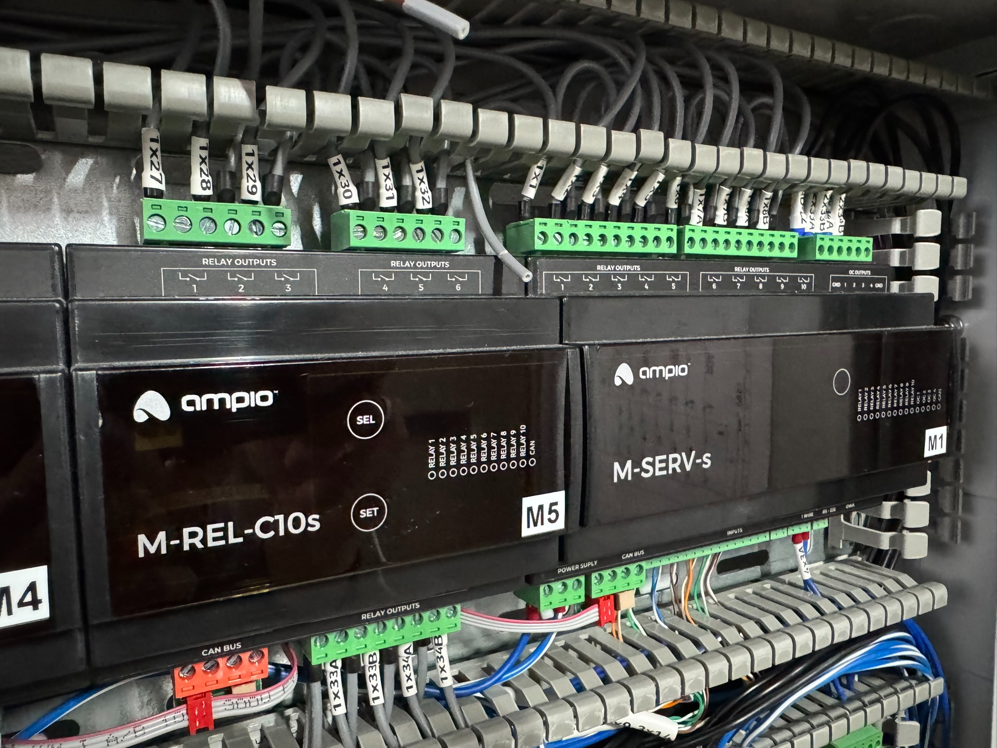 Rozdzielnica elektryczna z modułami sterującymi Ampio M-REL-C10s i M-SERV-s, widoczne liczne przewody, złącza śrubowe i oznaczenia.