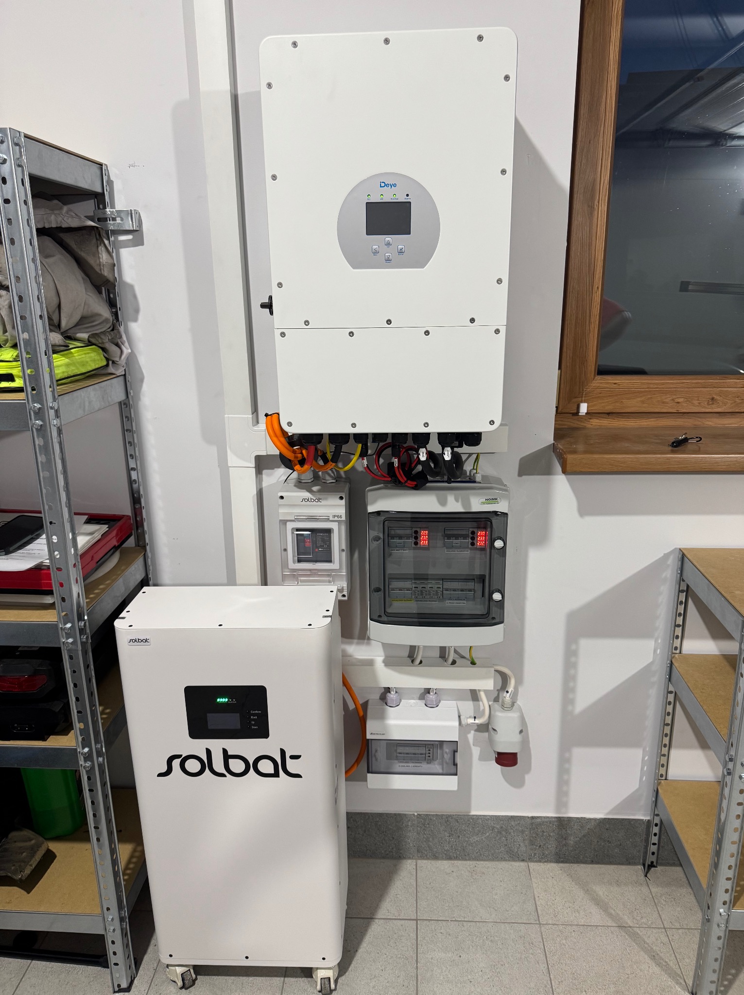 Instalacja magazynu energii Solbat z falownikiem Deye, widoczna rozdzielnica AC i zabezpieczenia MCCB. System zamontowany na ścianie, obok okna i metalowego regału.