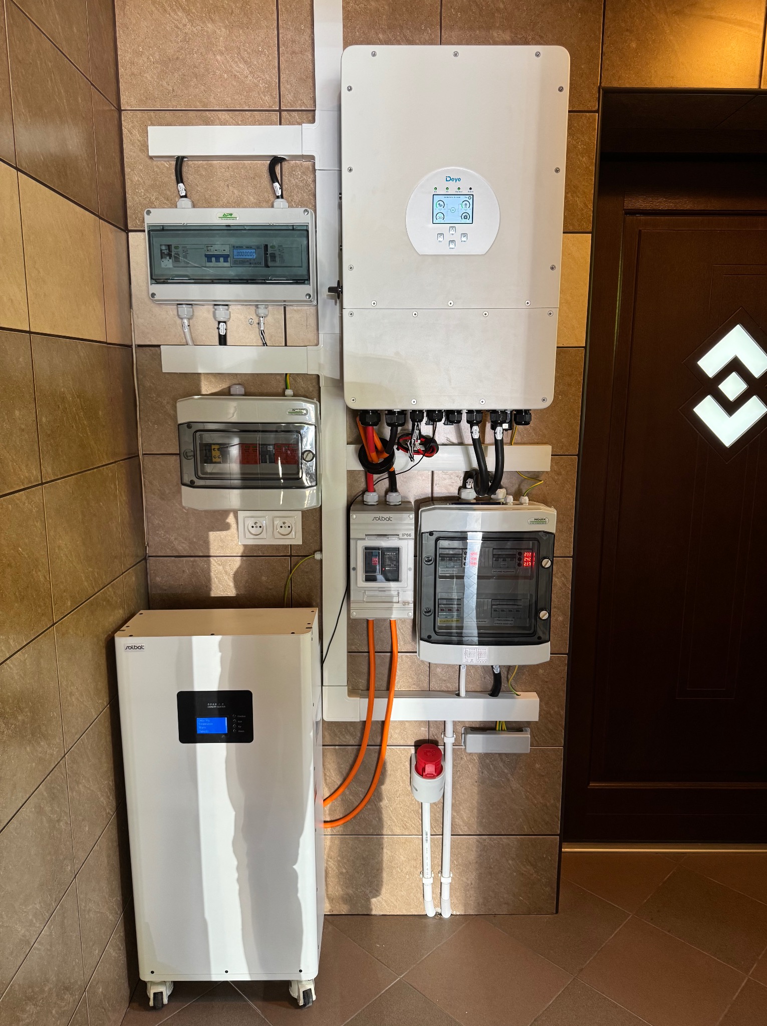 Zestaw magazynu energii Solbat z inwerterem Deye na ścianie z brązowych płytek. Widoczne skrzynki elektryczne i przewody w peszlach. Całość zamontowana obok ciemnych drzwi.