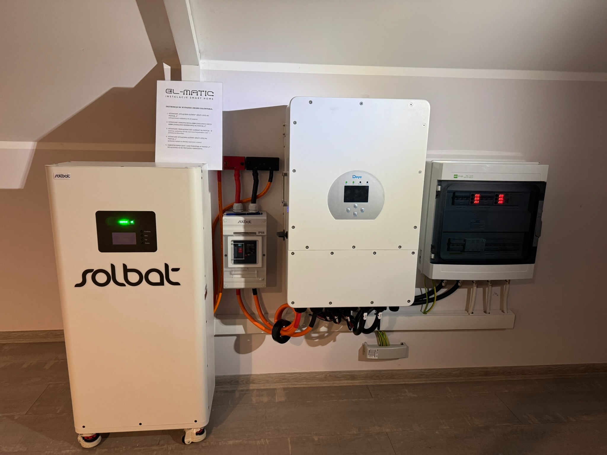 Instalacja fotowoltaiczna w pomieszczeniu: biały akumulator Solbat, inwerter Deye i rozdzielnica. Widoczne kable i instrukcja EL-MATIC.