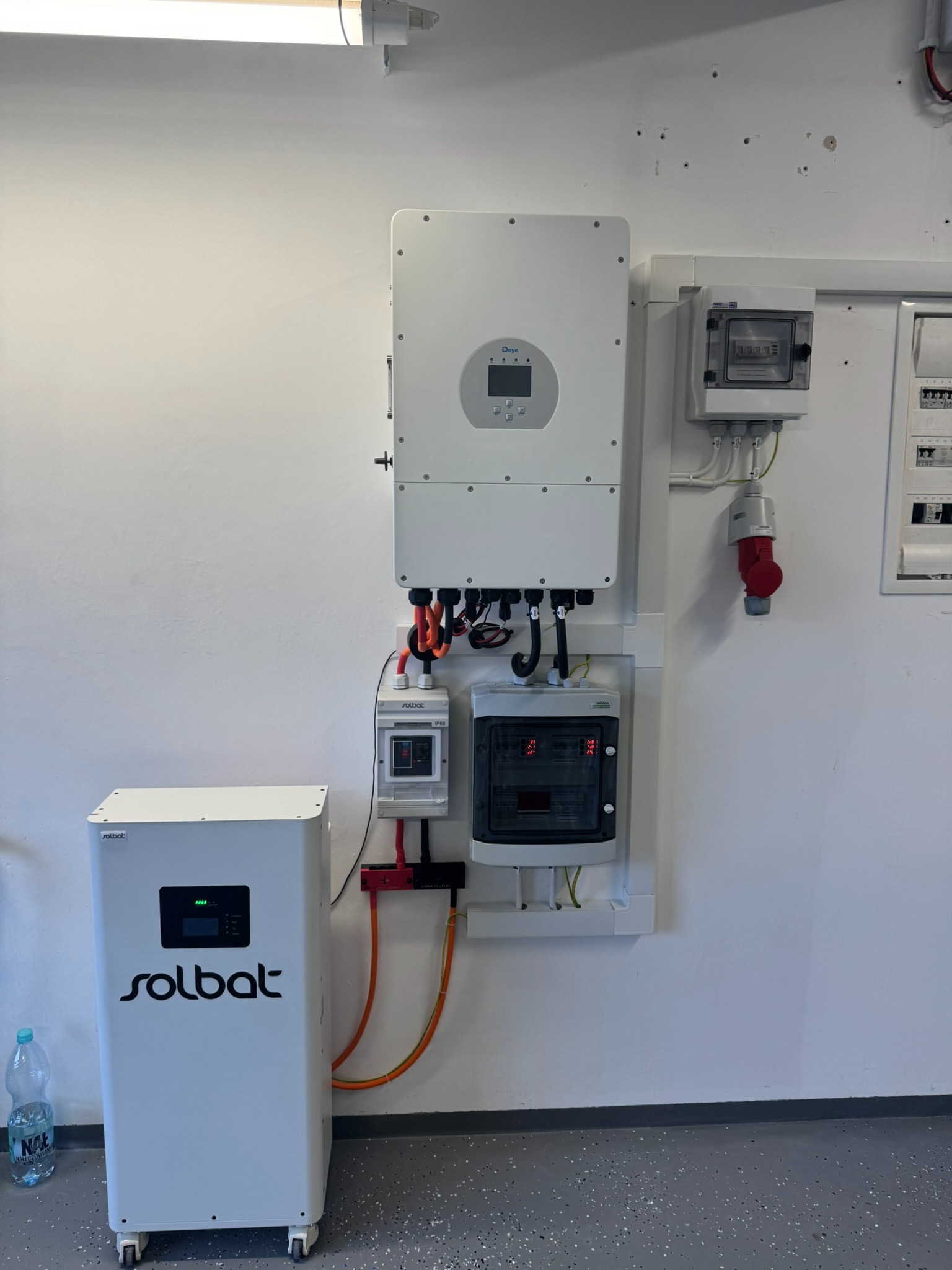 Biały magazyn energii Solbat z inwerterem Deye na ścianie, widoczne kable i skrzynki elektryczne. Instalacja w pomieszczeniu o jasnych ścianach i szarej podłodze.