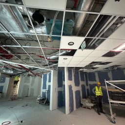 SK_Global - Montaż sufitu podwieszanego w biurze, widoczne instalacje nad sufitem, pracownik w żółtej kamizelce i kasku na rusztowaniu, ściany przygotowane pod malowanie z widocznymi łączeniami płyt