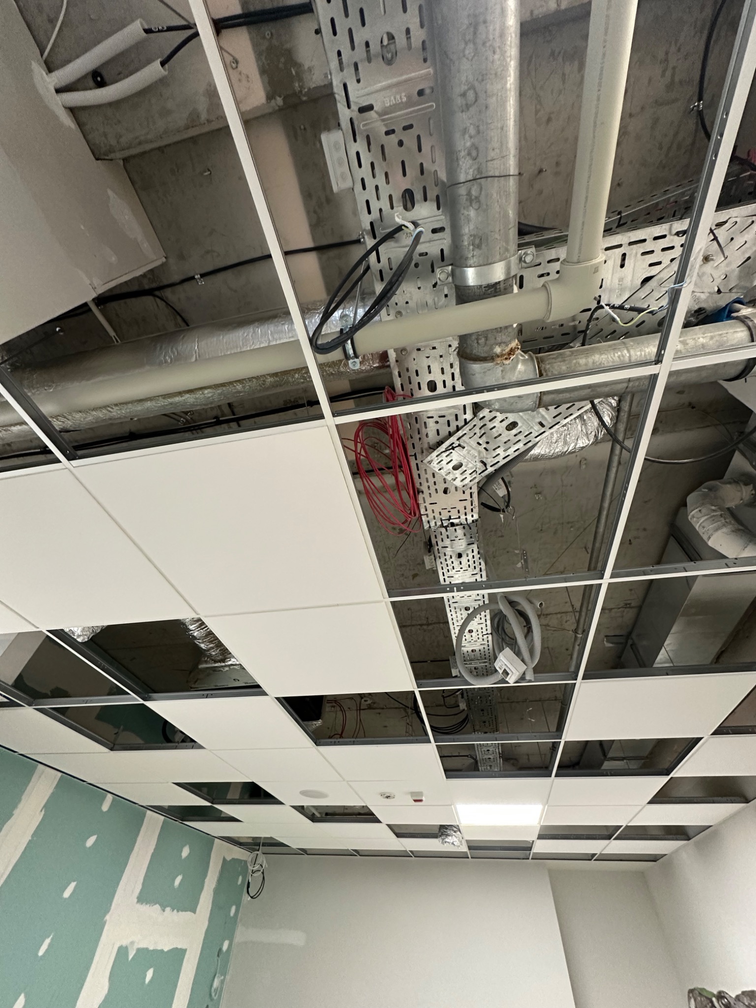 Sufit podwieszany z widoczną instalacją elektryczną i wentylacyjną, częściowo zakryty białymi panelami, ściana z płyt gipsowo-kartonowych w tle.