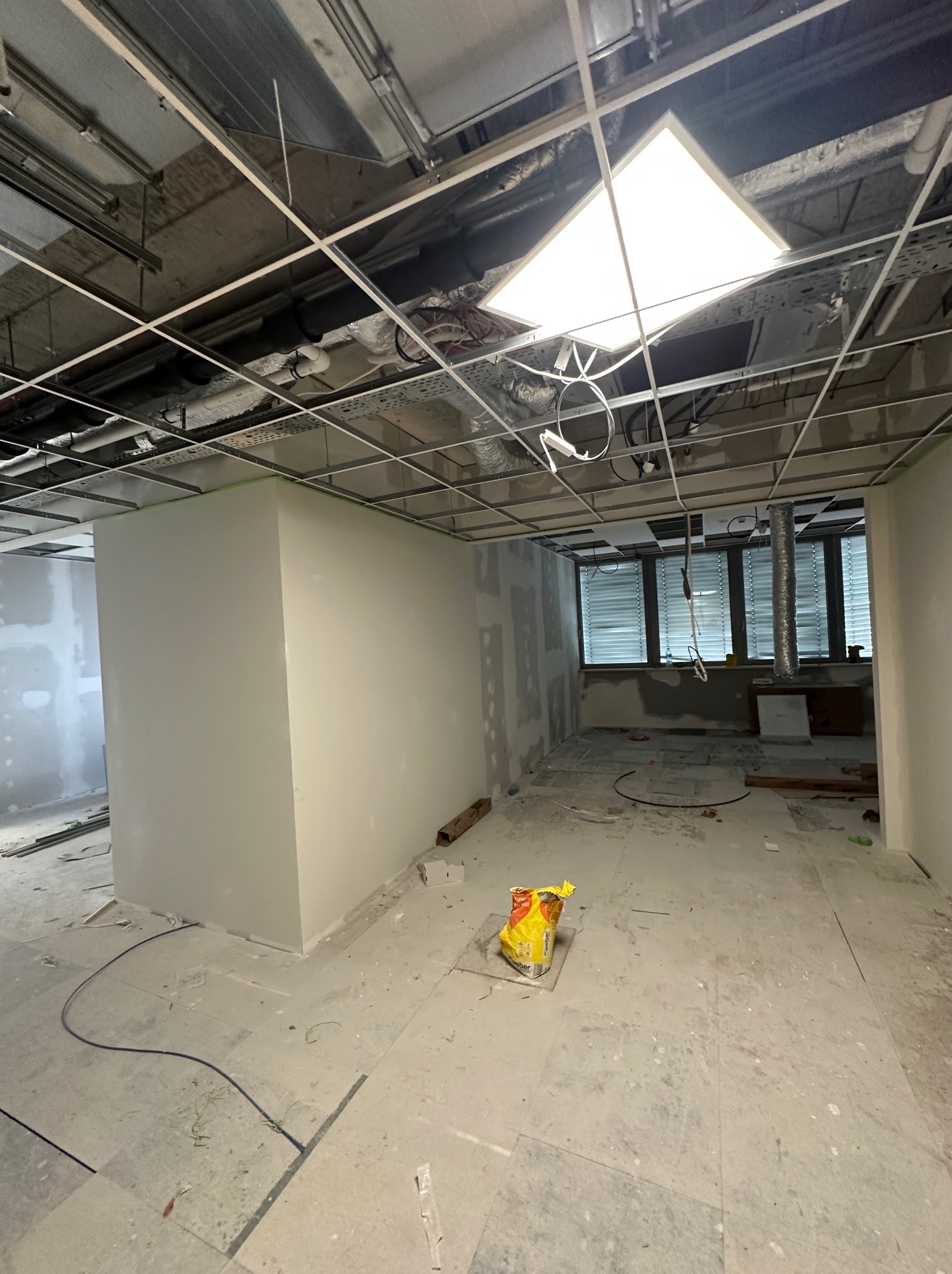 Wnętrze biura w trakcie remontu: ściany po gładziowaniu, widoczne instalacje pod sufitem, worek z zaprawą na podłodze, okna z żaluzjami w tle.