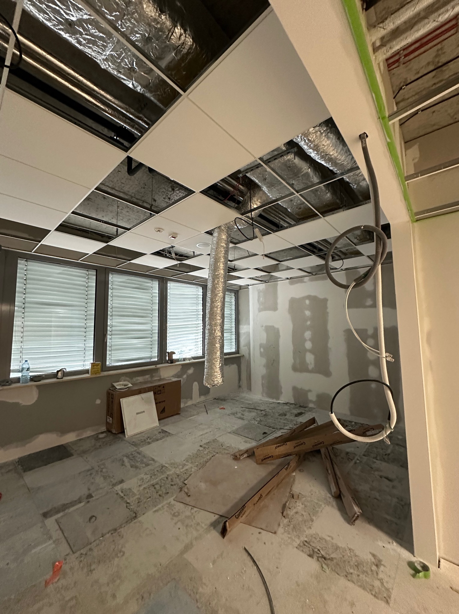 Wnętrze biura w trakcie prac wykończeniowych: widoczne podwieszane sufity, surowe ściany przygotowane do malowania, instalacje elektryczne i wentylacyjne, oraz materiały budowlane na podłodze.