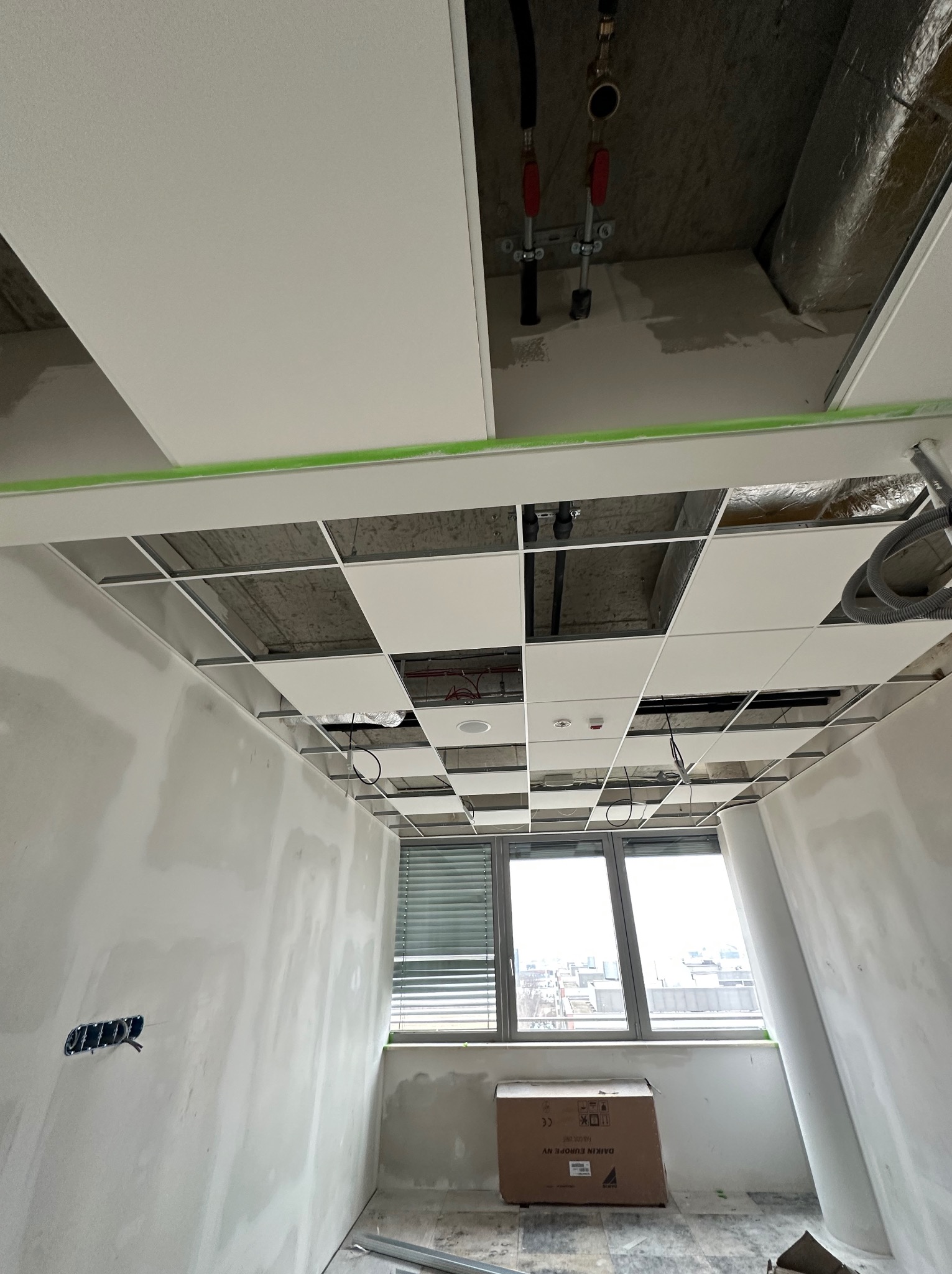 Wnętrze biura w trakcie prac wykończeniowych, widoczny sufit podwieszany z częściowo zamontowanymi panelami, ściany po szpachlowaniu, okno z widokiem na miasto, karton na podłodze.