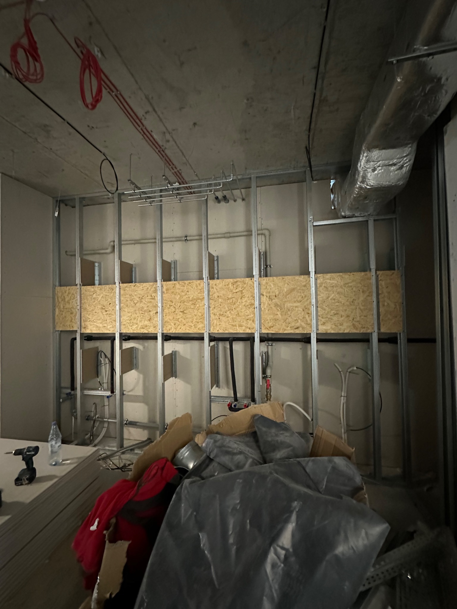 Stelaż metalowy pod zabudowę w surowym stanie, widoczne instalacje, płyty OSB w konstrukcji, przewody elektryczne pod sufitem, narzędzia i materiały budowlane w nieładzie na podłodze.