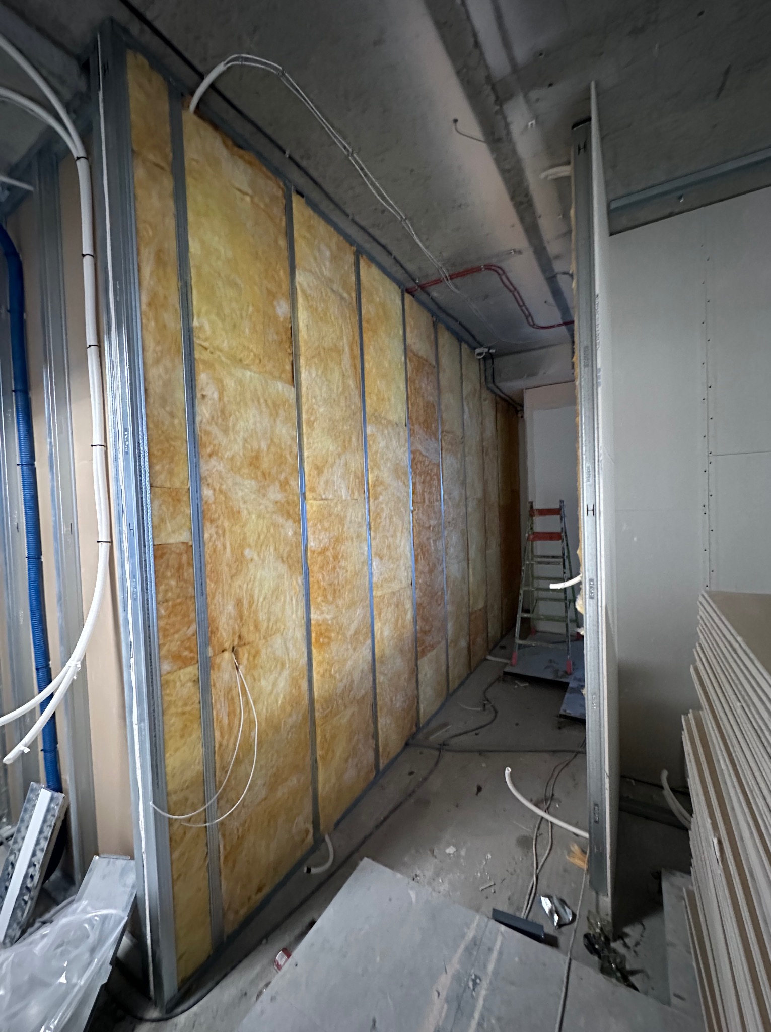 Montaż izolacji z wełny mineralnej w ścianie szkieletowej z profili stalowych, widoczne przewody elektryczne i rury instalacyjne, w tle drabina i płyty gipsowo-kartonowe.