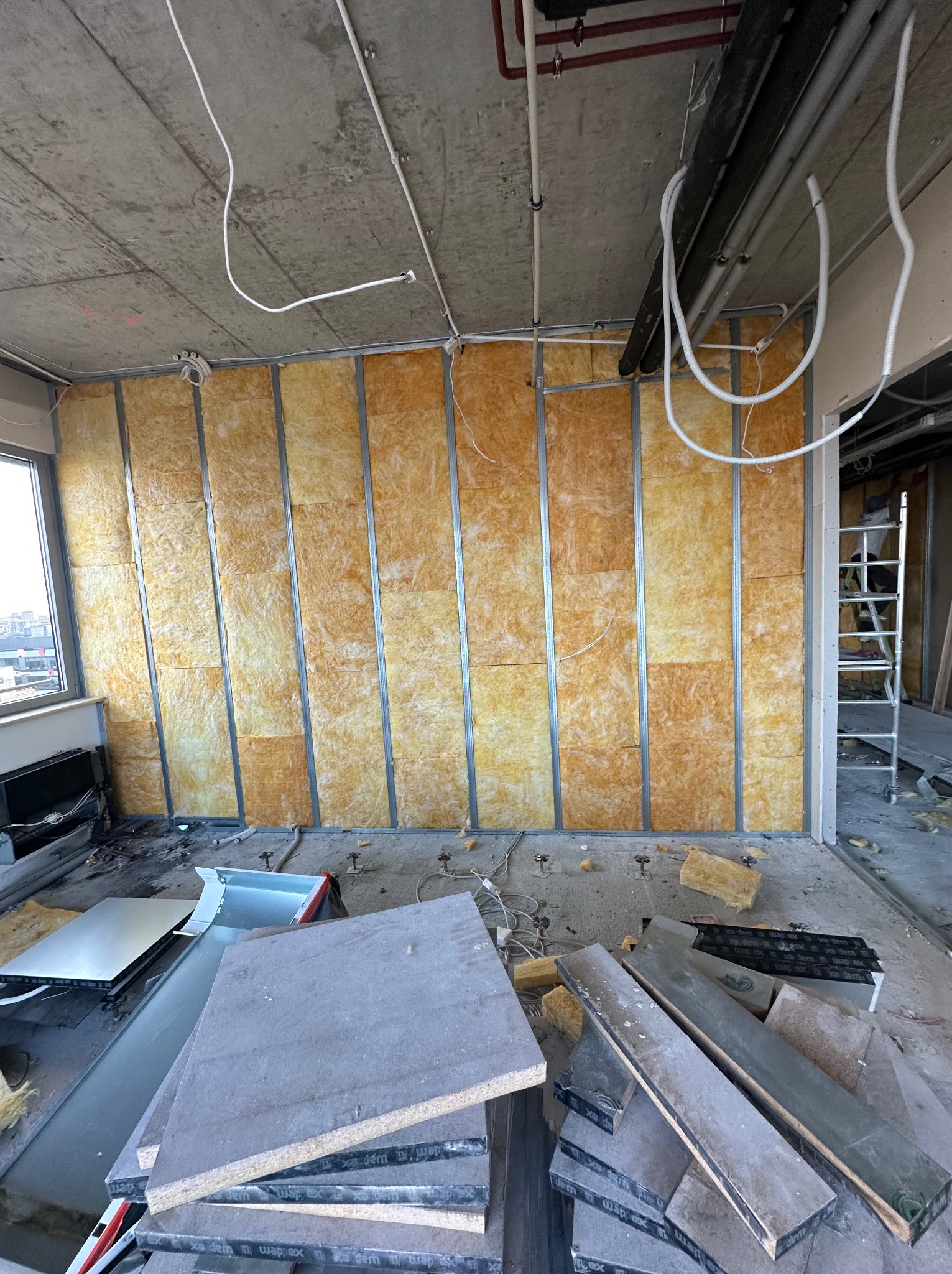 Montaż izolacji z wełny mineralnej w metalowym stelażu na ścianie w surowym stanie, widoczne przewody, elementy instalacji pod sufitem i stos płyt na podłodze.