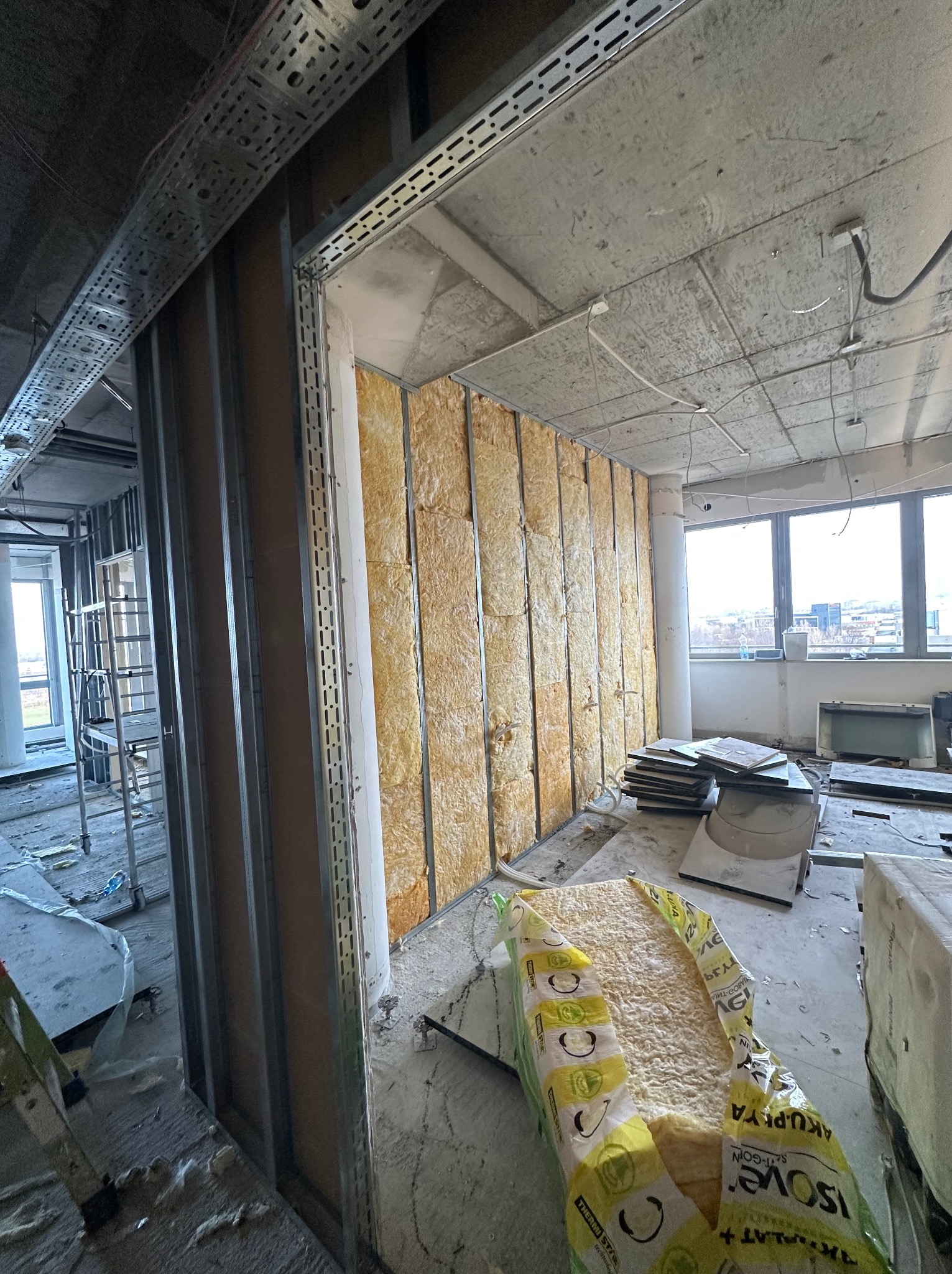 Montaż izolacji z wełny mineralnej w metalowym stelażu ściany działowej w surowym wnętrzu biurowym z widocznymi instalacjami pod sufitem i dużymi oknami w tle.