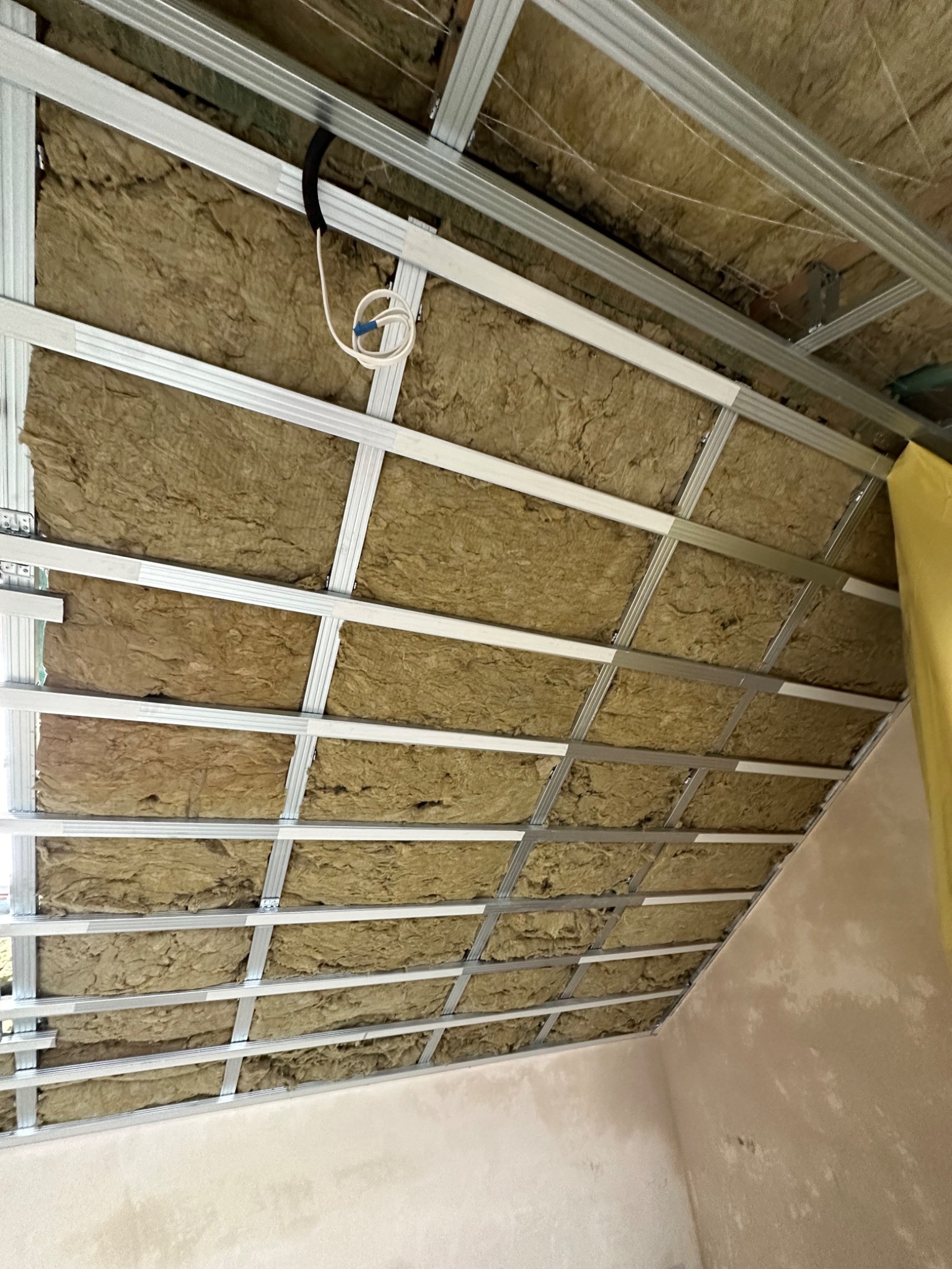 Sufit z widoczną konstrukcją metalową i izolacją z wełny mineralnej, przewody elektryczne zwisają z sufitu, ściana w tle przygotowana pod malowanie.