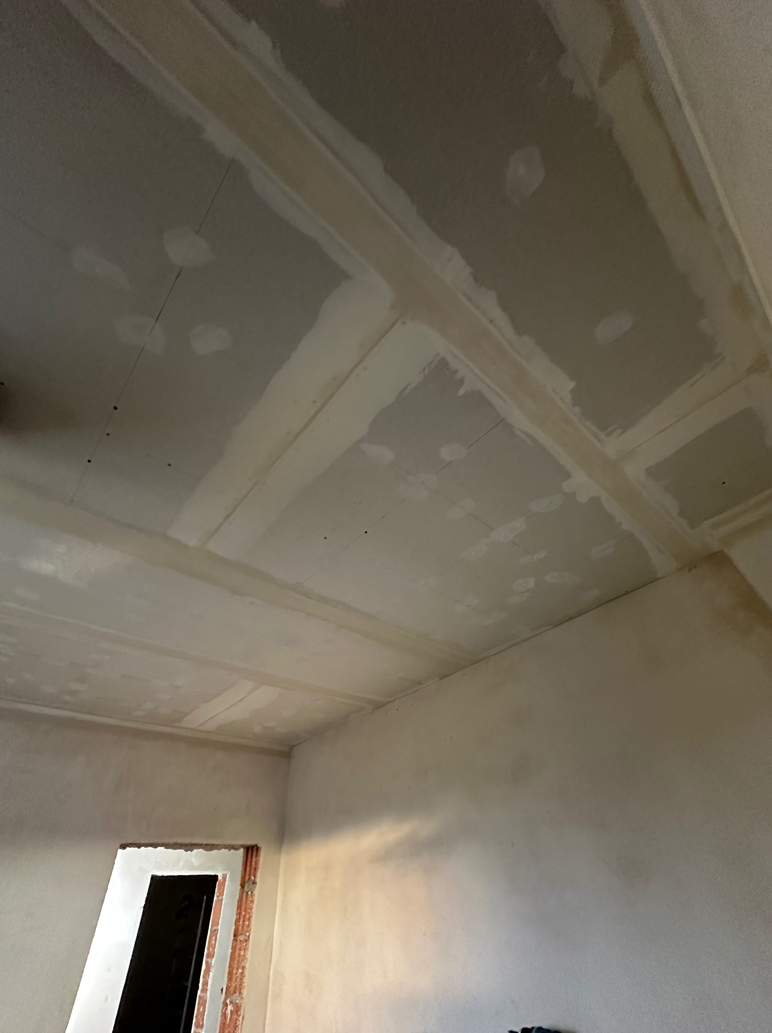Sufit z płyt kartonowo-gipsowych w trakcie szpachlowania łączeń, widoczne ślady po wkrętach, surowy stan wykończenia pomieszczenia z widocznym otworem drzwiowym.