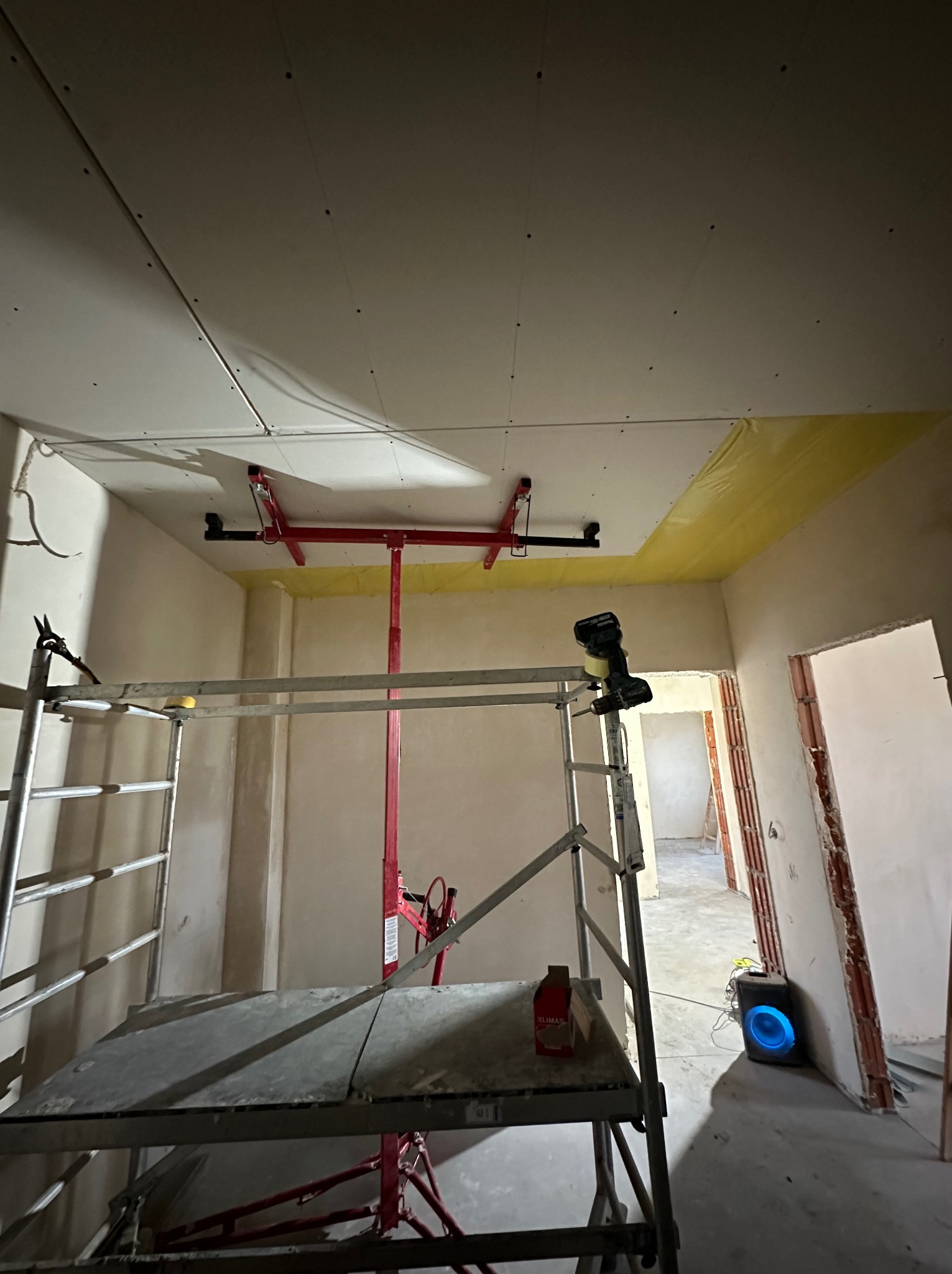 Montaż płyt gipsowo-kartonowych na suficie przy użyciu windy, widoczny rusztowanie, żółta folia zabezpieczająca ścianę, narzędzia i głośnik w tle.
