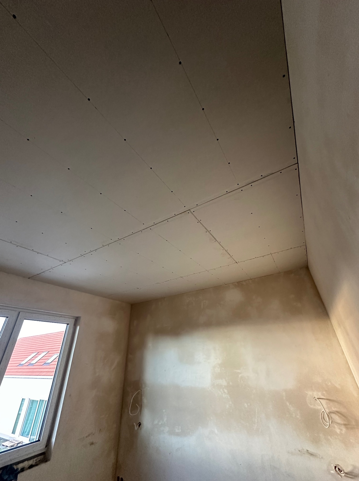 Montaż płyt karton gips na suficie w pomieszczeniu z oknem, widoczne łączenia płyt, ślady po szpachlowaniu na ścianach, przygotowanie do dalszych prac wykończeniowych.