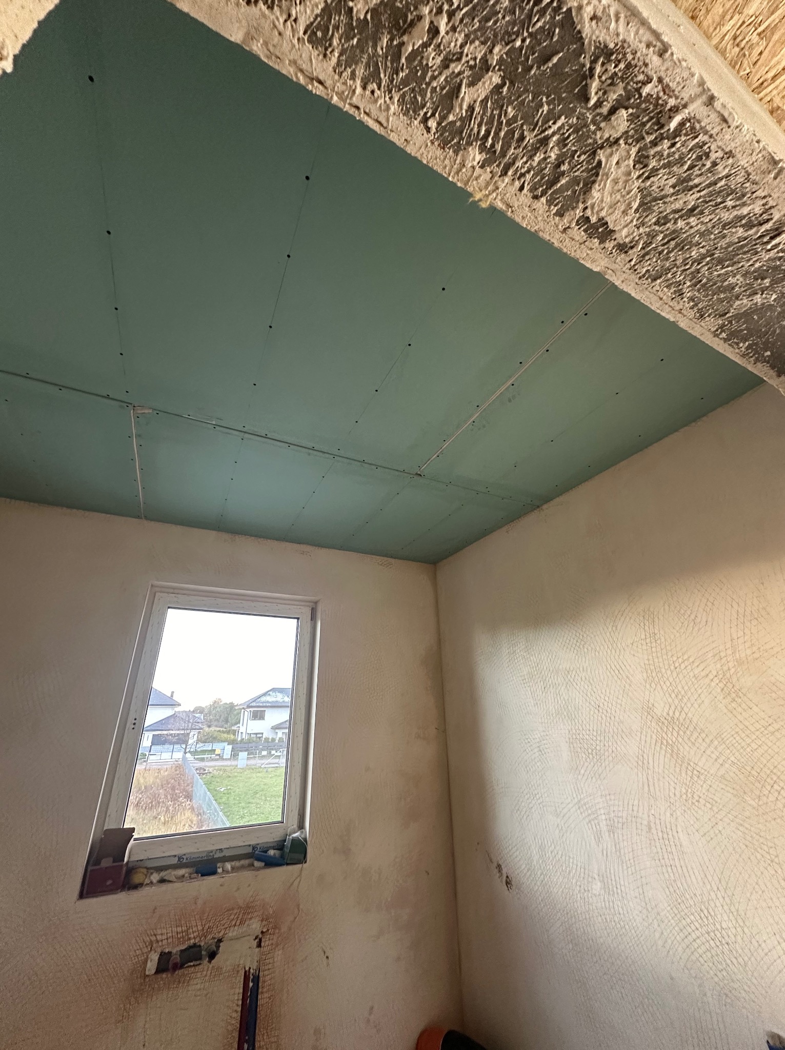 Wnętrze w trakcie prac wykończeniowych: sufit z zielonych płyt, ściany przygotowane do malowania z widoczną fakturą, okno z widokiem na zewnątrz.