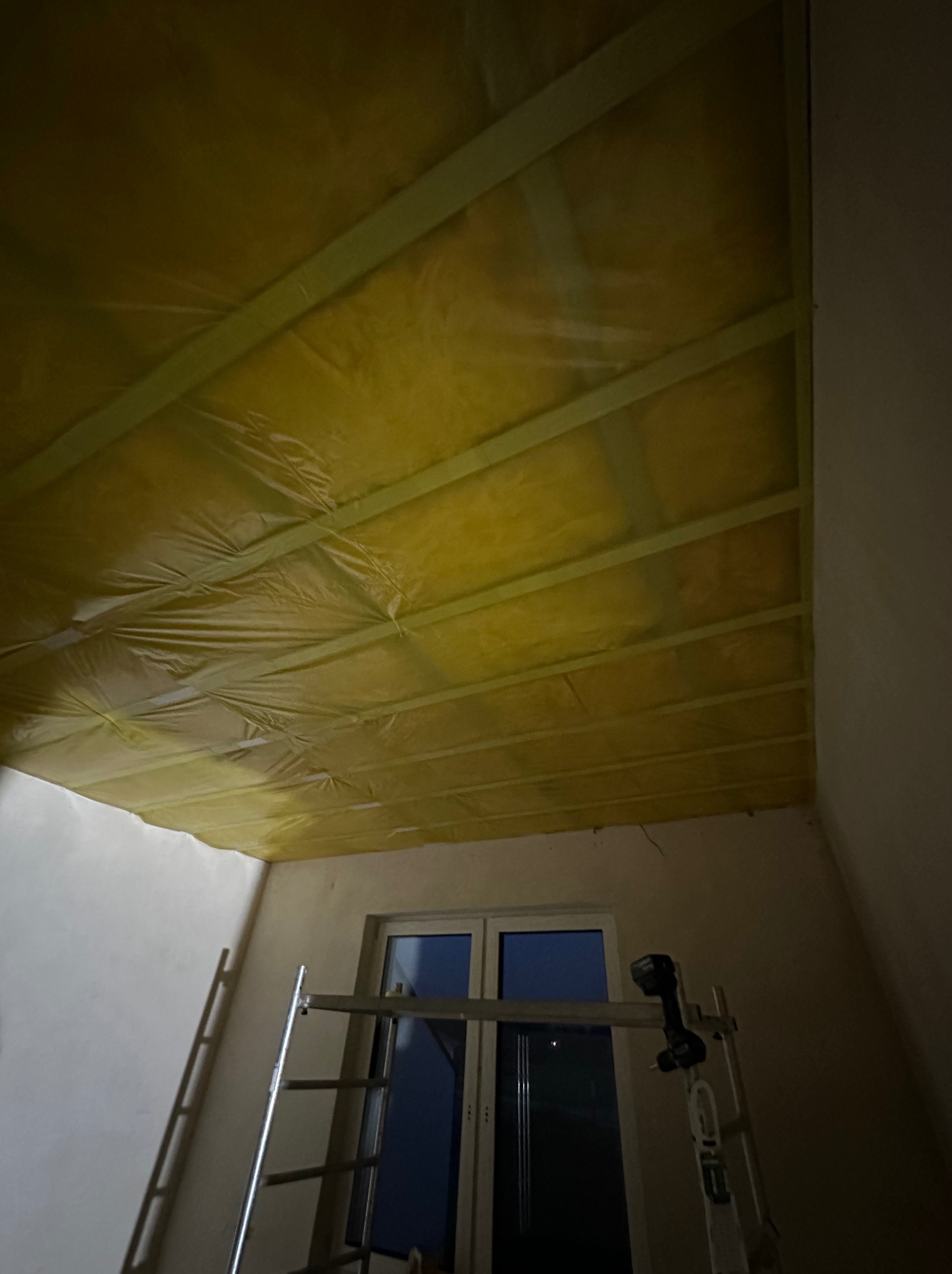 Żółta wełna mineralna zabezpieczona folią zamontowana między krokwiami na poddaszu, widoczne rusztowanie i drabina oparta o ścianę przy oknie.