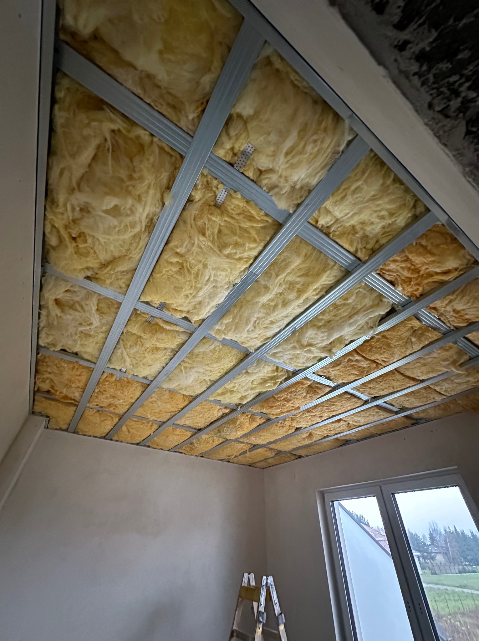 Montaż izolacji z wełny mineralnej na poddaszu, widoczna metalowa konstrukcja nośna sufitu podwieszanego, drabina oparta o ścianę, okno dachowe w tle.