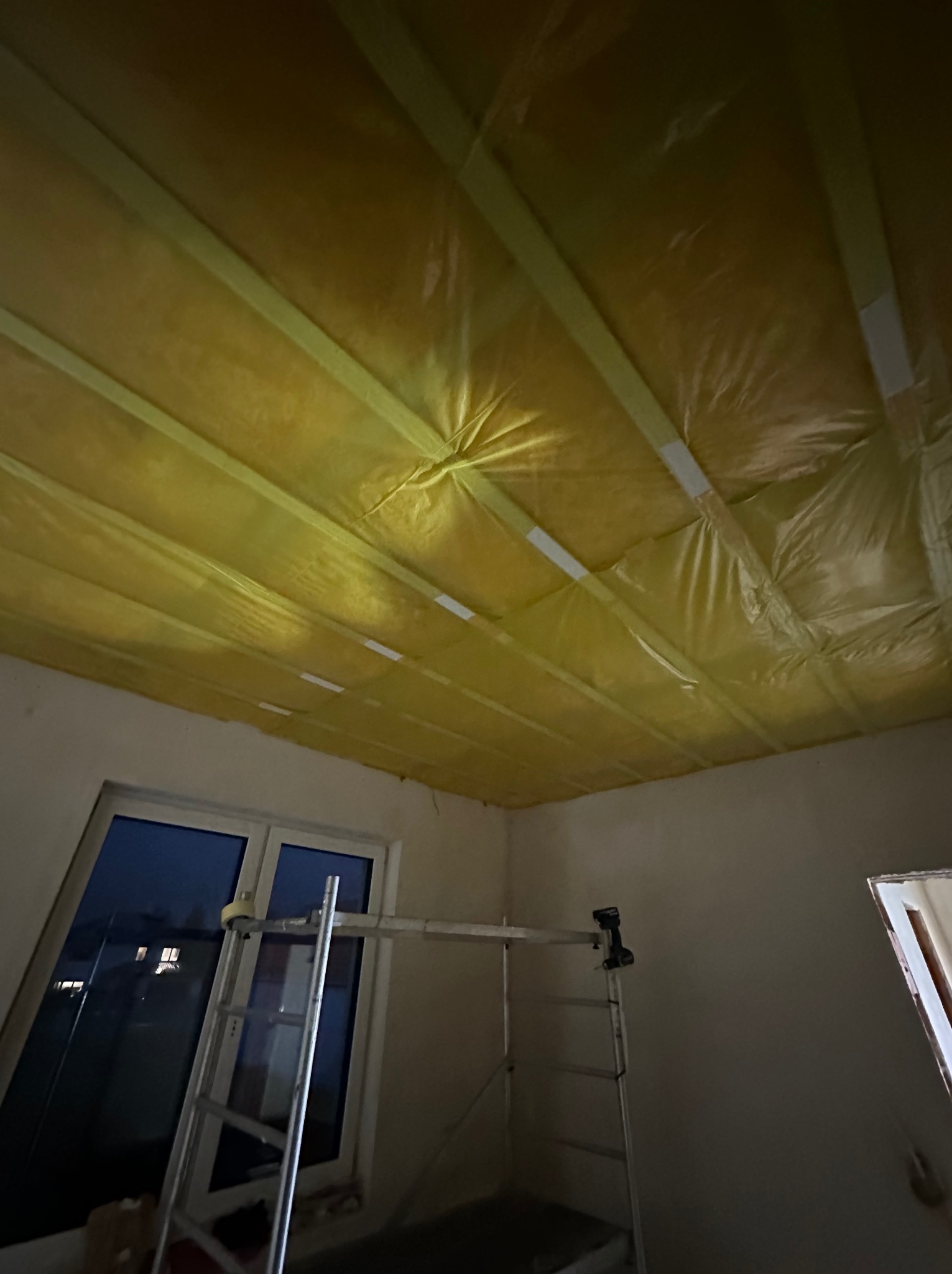 Przygotowanie do ocieplenia poddasza w Warszawie: widok na sufit z folią paroizolacyjną, rusztowanie w pomieszczeniu, okno w tle.