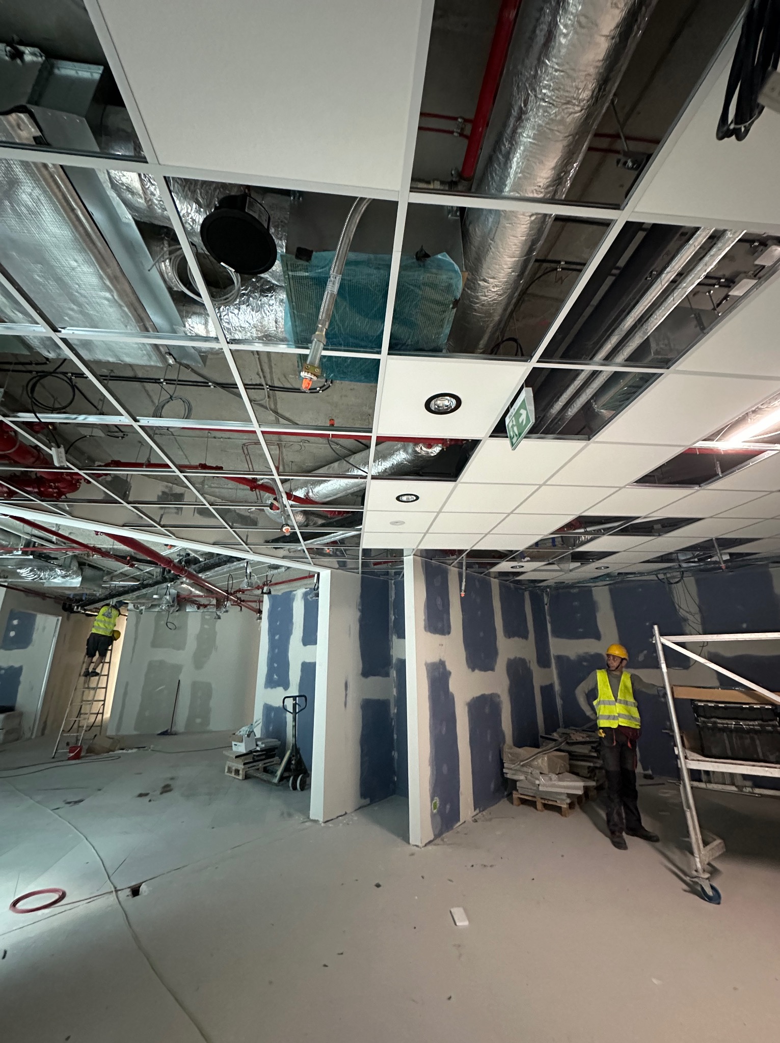 Montaż sufitu podwieszanego w biurze, widoczne instalacje nad sufitem, pracownik w żółtej kamizelce i kasku na rusztowaniu, ściany przygotowane pod malowanie z widocznymi łączeniami płyt