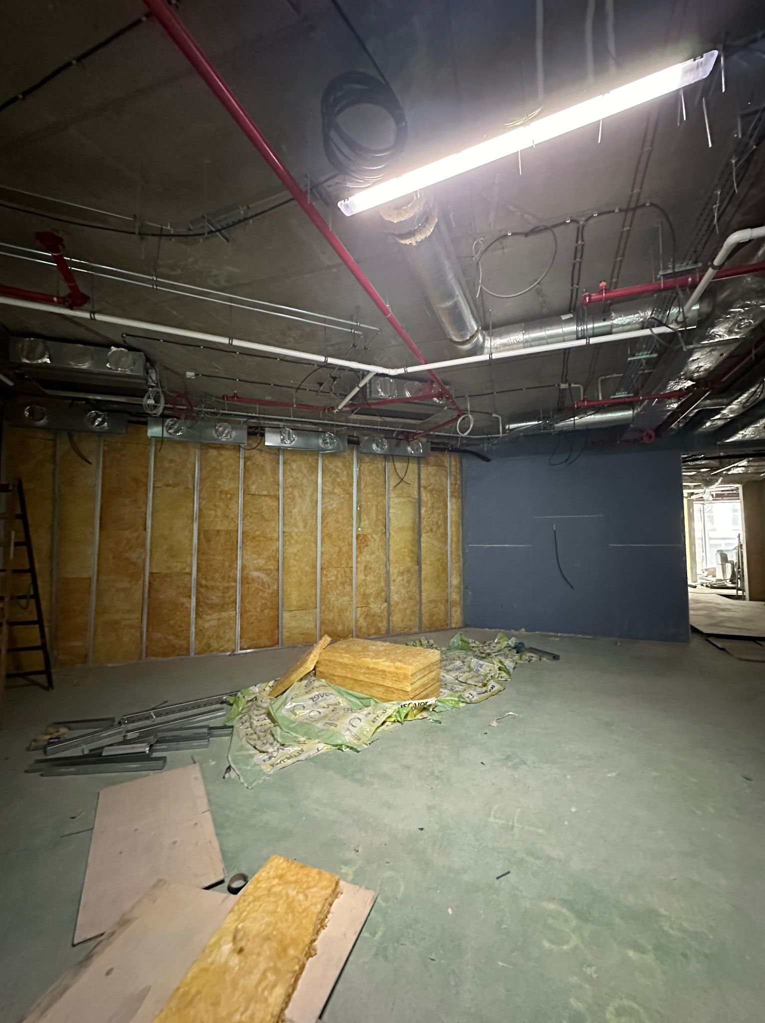 Montaż izolacji z wełny mineralnej na ścianie z profili stalowych w surowym wnętrzu biurowym, widoczne instalacje pod sufitem i narzędzia pracy.