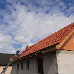 Pracownicy na drabinie układają czerwoną dachówkę na nowym dachu domu w stanie surowym, widoczne niebieskie niebo z białymi chmurami.
