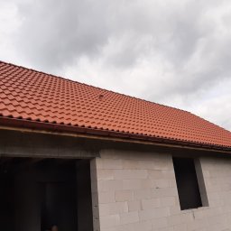 Osoba prywatna - Nowy, czerwony dach z dachówki ceramicznej na budynku w trakcie budowy, widoczne białe bloczki betonowe i zamontowane rynny w kolorze dachu, pochmurne niebo w tle.