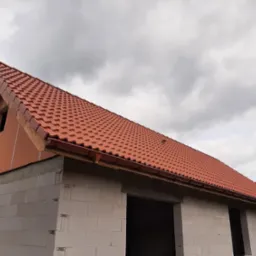 Czerwona dachówka ceramiczna na nowym domu w trakcie budowy, widoczne rynny i ściany z betonu komórkowego pod pochmurnym niebem.