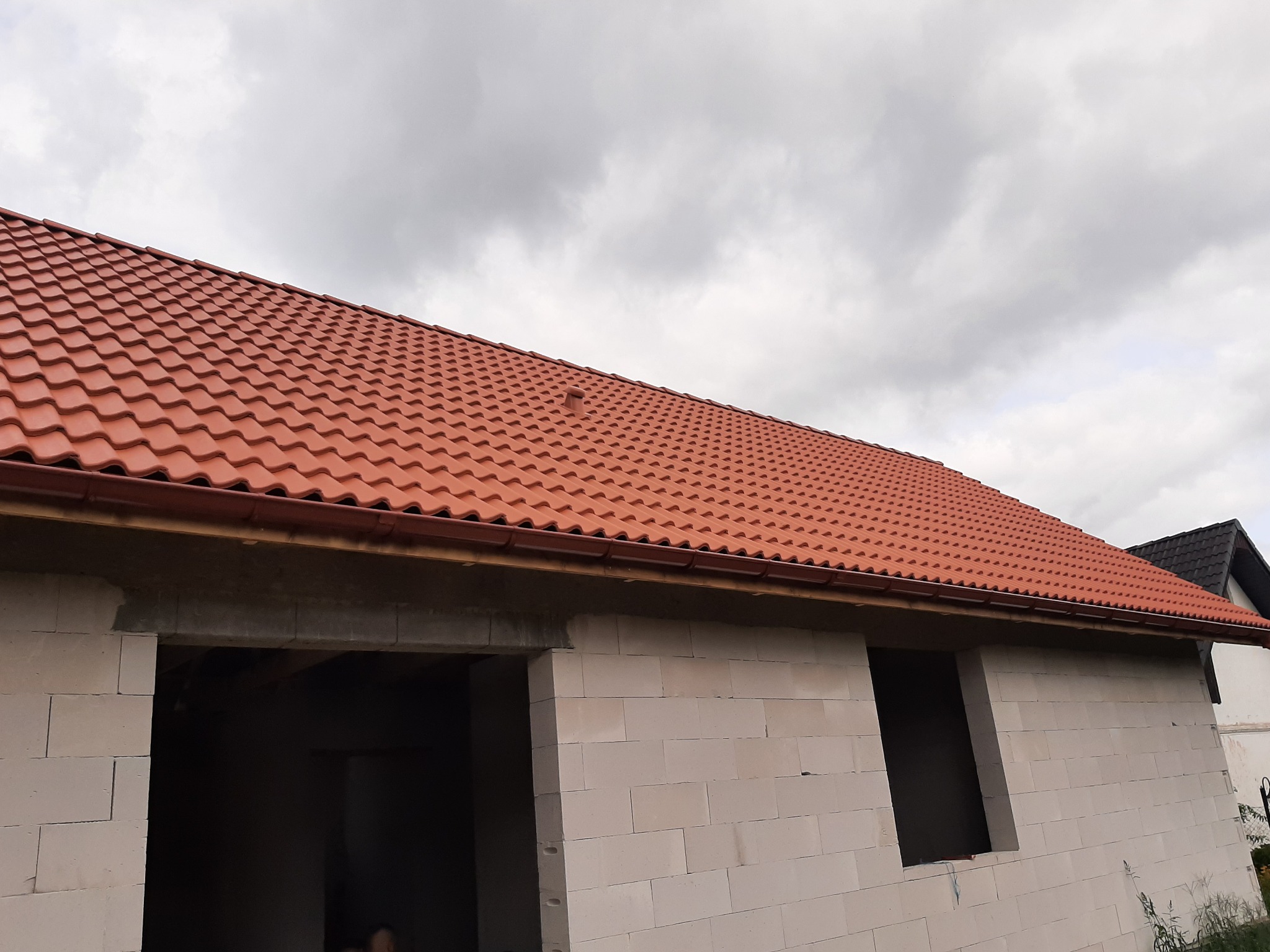 Nowy, czerwony dach z dachówki ceramicznej na budynku w trakcie budowy, widoczne białe bloczki betonowe i zamontowane rynny w kolorze dachu, pochmurne niebo w tle.