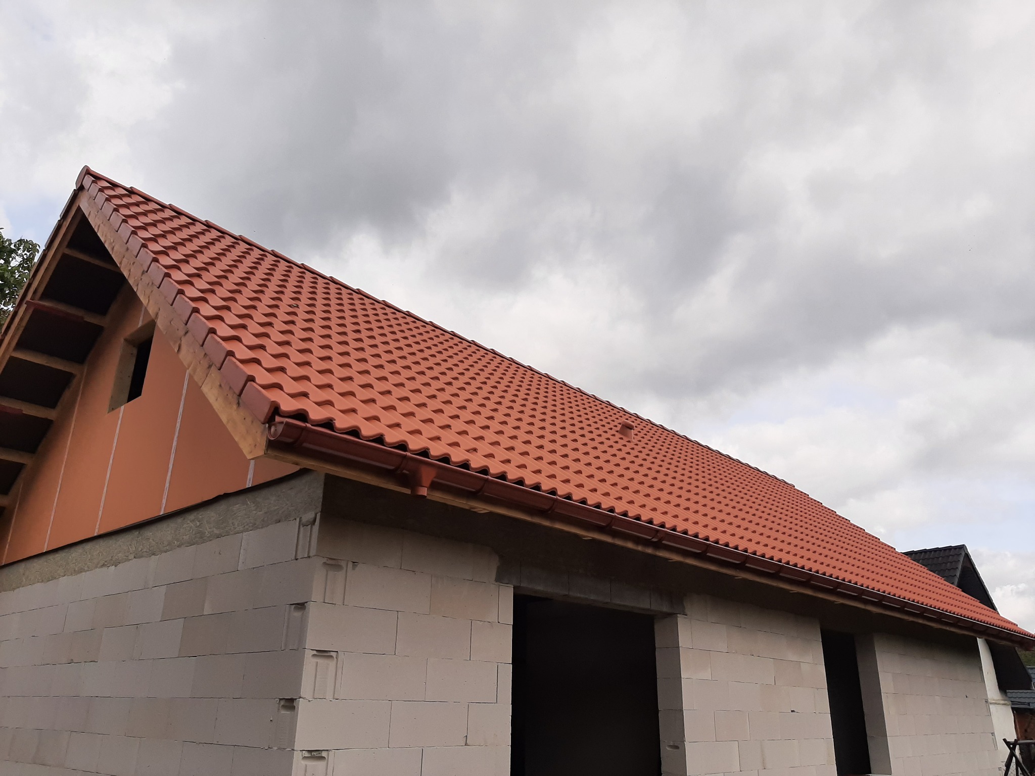 Czerwona dachówka ceramiczna na nowym domu w trakcie budowy, widoczne rynny i ściany z betonu komórkowego pod pochmurnym niebem.