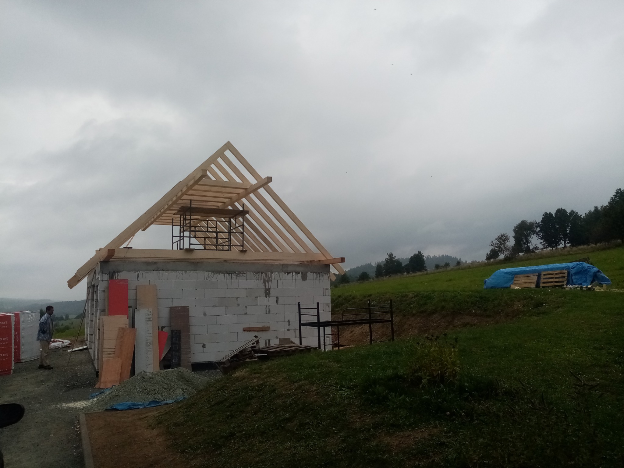 Surowa konstrukcja domu z bloczków, widoczna drewniana więźba dachowa w trakcie budowy, materiały budowlane składowane obok, krajobraz pagórkowaty w tle, pochmurne niebo.