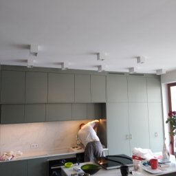 ELECTRO SERVICE SPÓŁKA Z OGRANICZONĄ ODPOWIEDZIALNOŚCIĄ - Montaż oświetlenia sufitowego w kuchni: elektryk pochylony nad blatem, instaluje białe, kwadratowe lampy w nowoczesnej zabudowie kuchennej w odcieniach szarości i bieli.