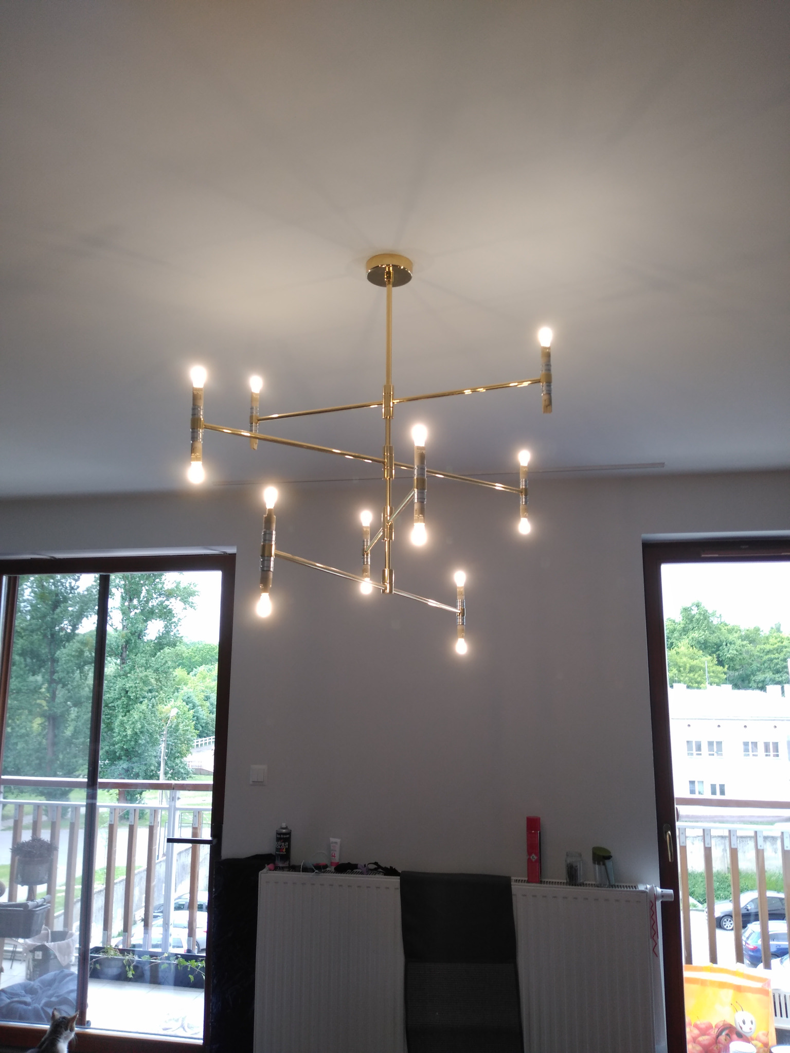 Nowoczesny, złoty żyrandol z wieloma ramionami i żarówkami, zamontowany w jasnym pokoju z oknami balkonowymi. Widok na zewnątrz, na zieleń i budynki.