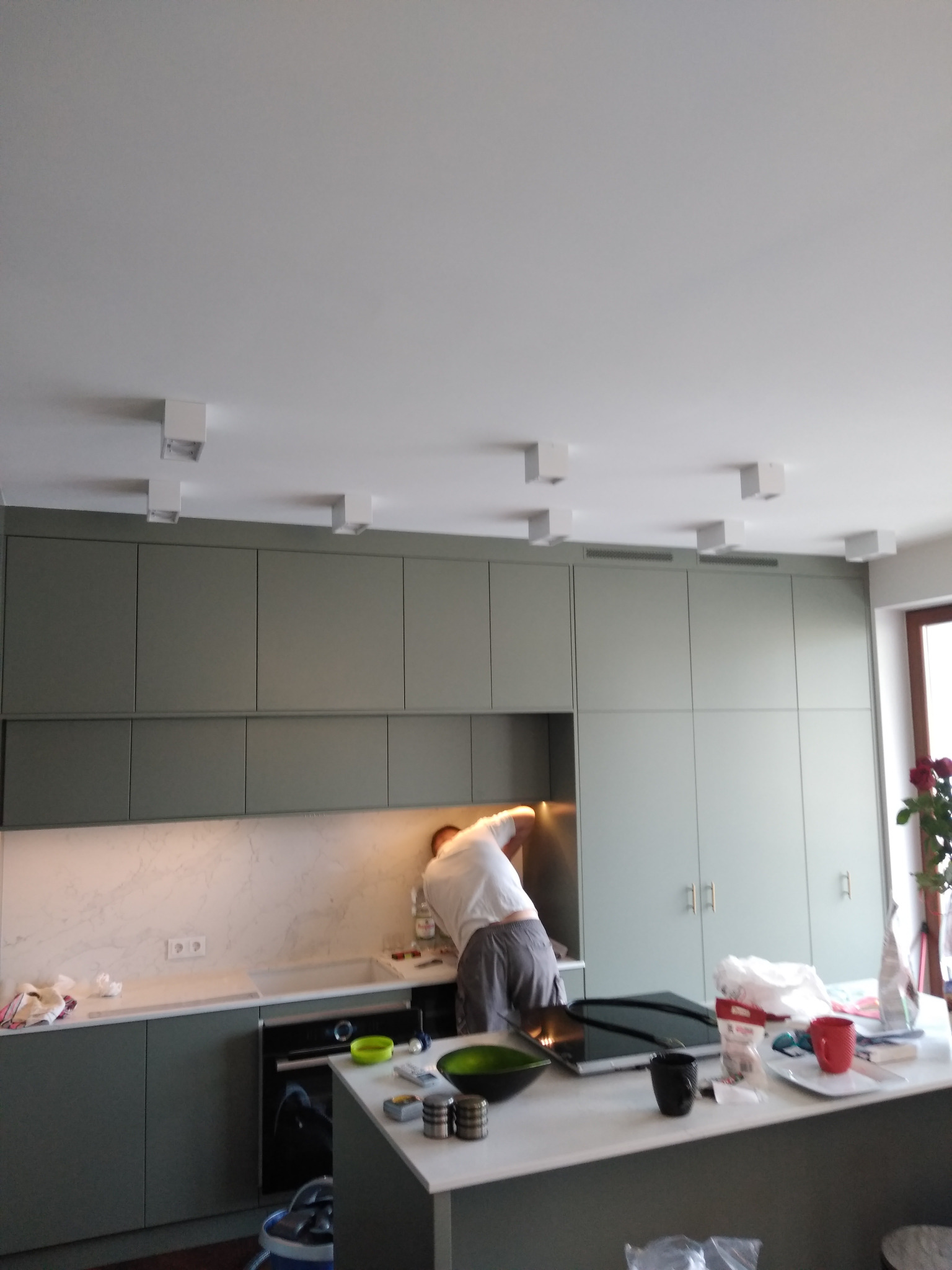 Montaż oświetlenia sufitowego w kuchni: elektryk pochylony nad blatem, instaluje białe, kwadratowe lampy w nowoczesnej zabudowie kuchennej w odcieniach szarości i bieli.