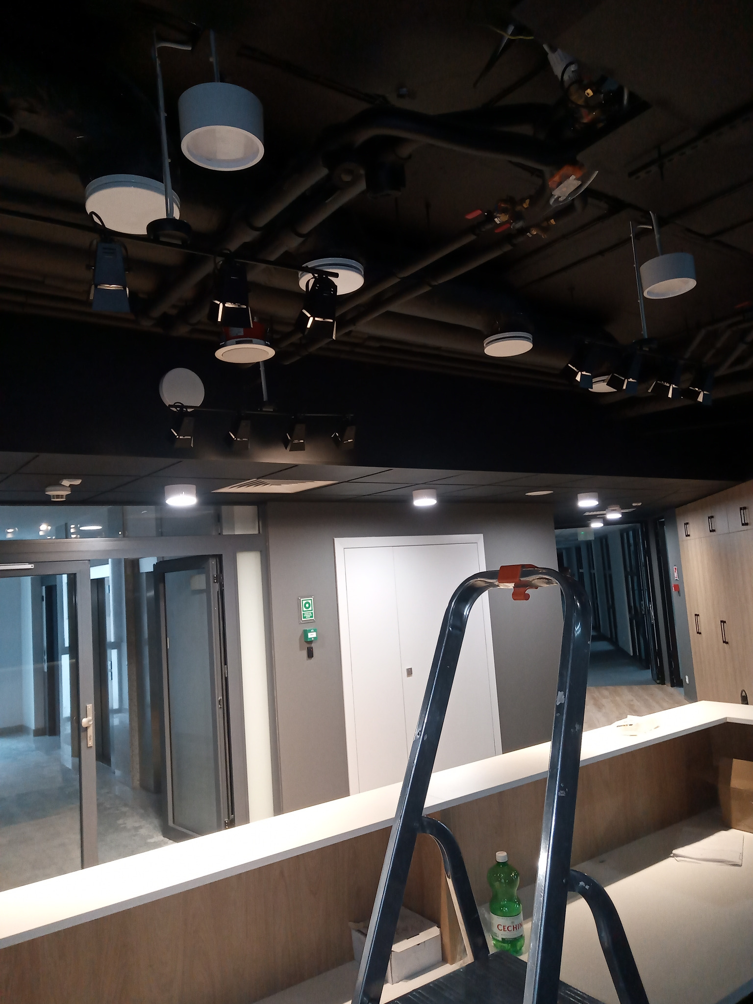 Montaż oświetlenia sufitowego w biurze: widoczne lampy, rury instalacyjne pod sufitem, drabina i butelka. Ujęcie z dołu, w trakcie prac.