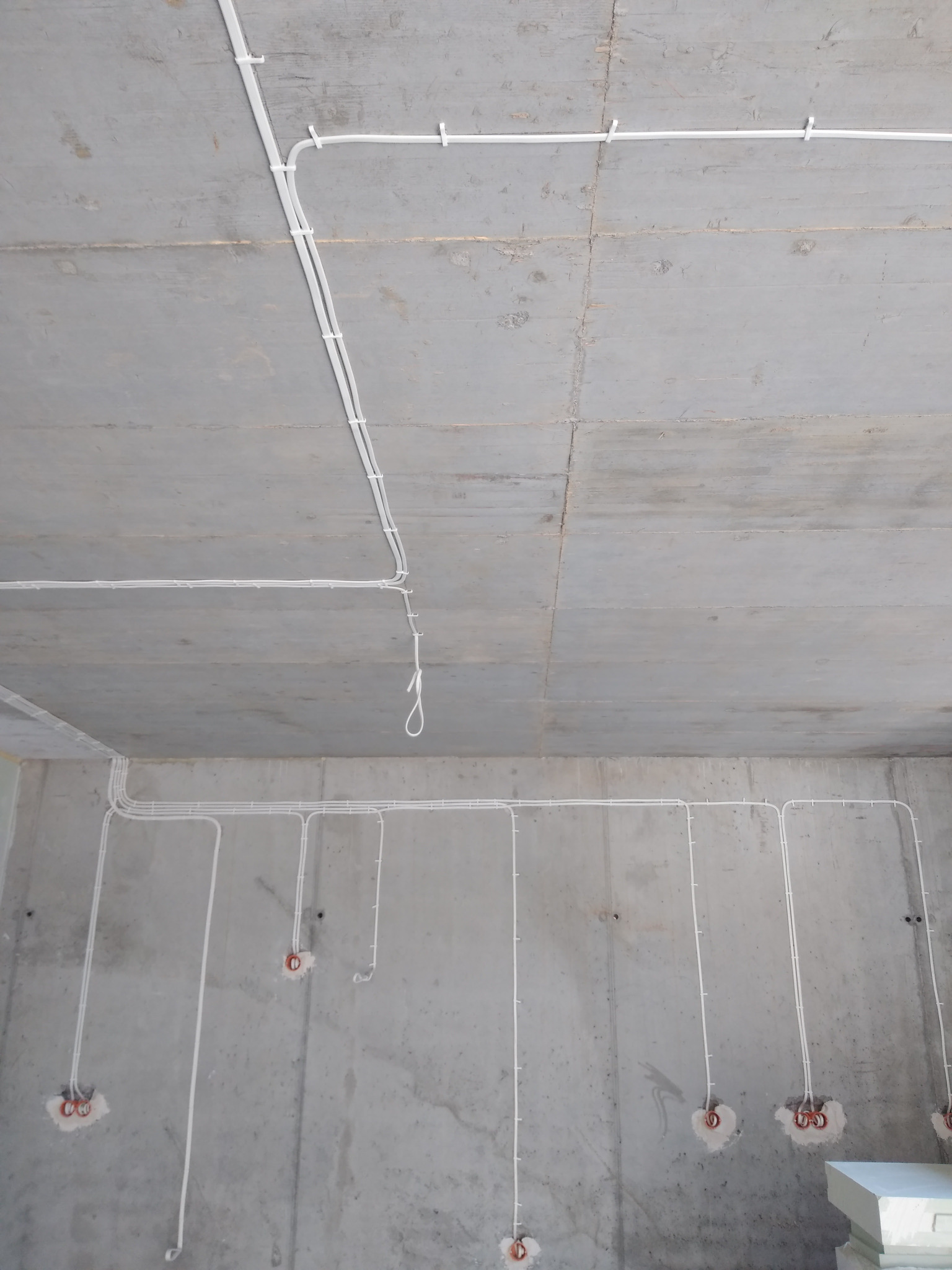 Białe kable elektryczne prowadzone po surowym, betonowym suficie i ścianach w nowym budynku. Widoczne puszki instalacyjne w ścianach. Instalacja elektryczna w trakcie montażu.