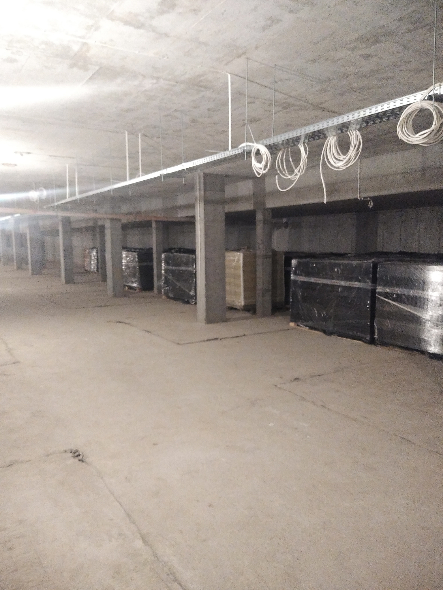 Surowy strop z korytkami kablowymi i zwisającymi kablami w nowym budynku. W tle materiały budowlane pod folią.