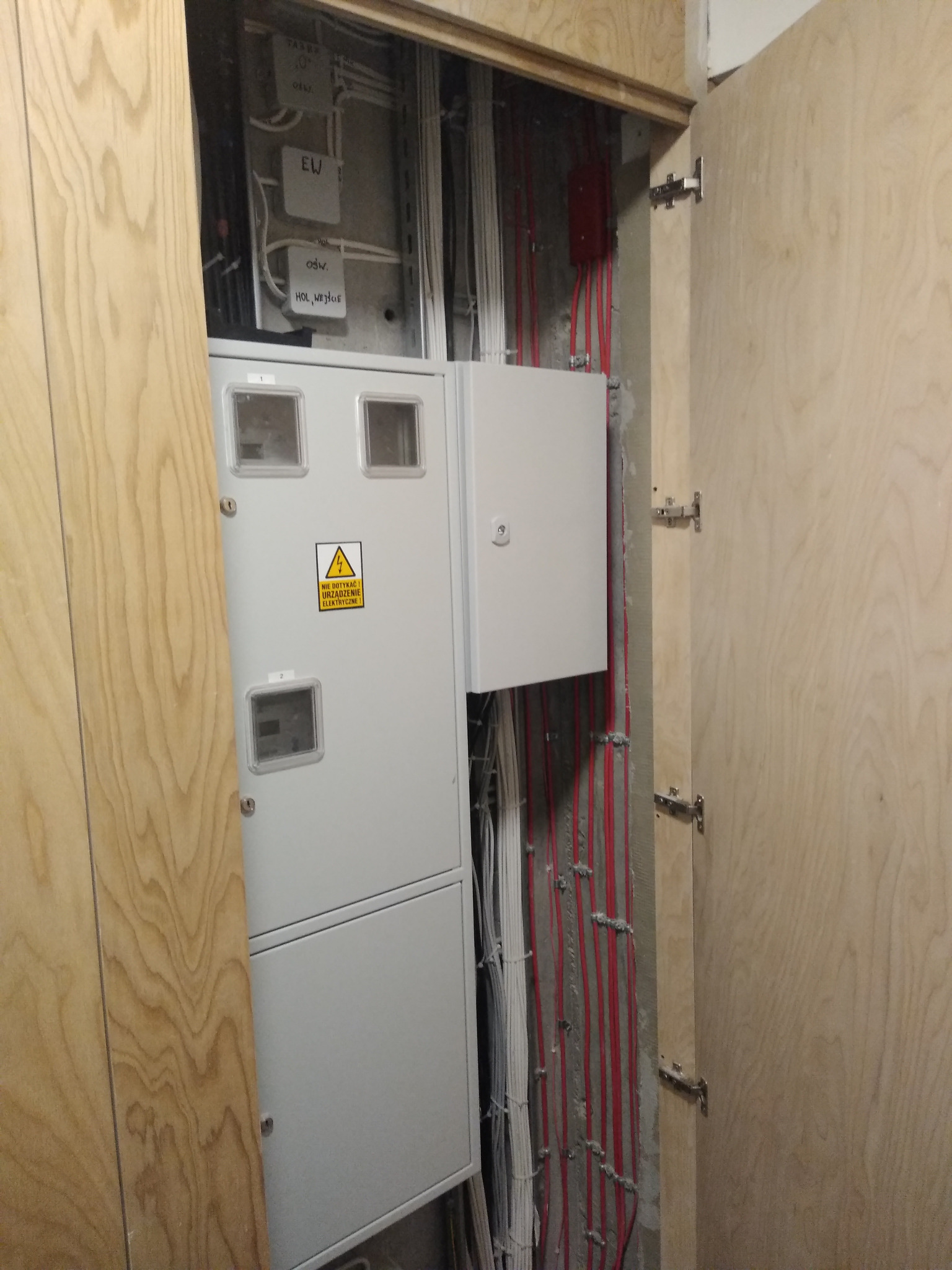 Szara szafka elektryczna z żółtą naklejką ostrzegawczą 'Nie dotykać! Urządzenie elektryczne!' w drewnianej wnęce z widocznymi kablami w ścianie.