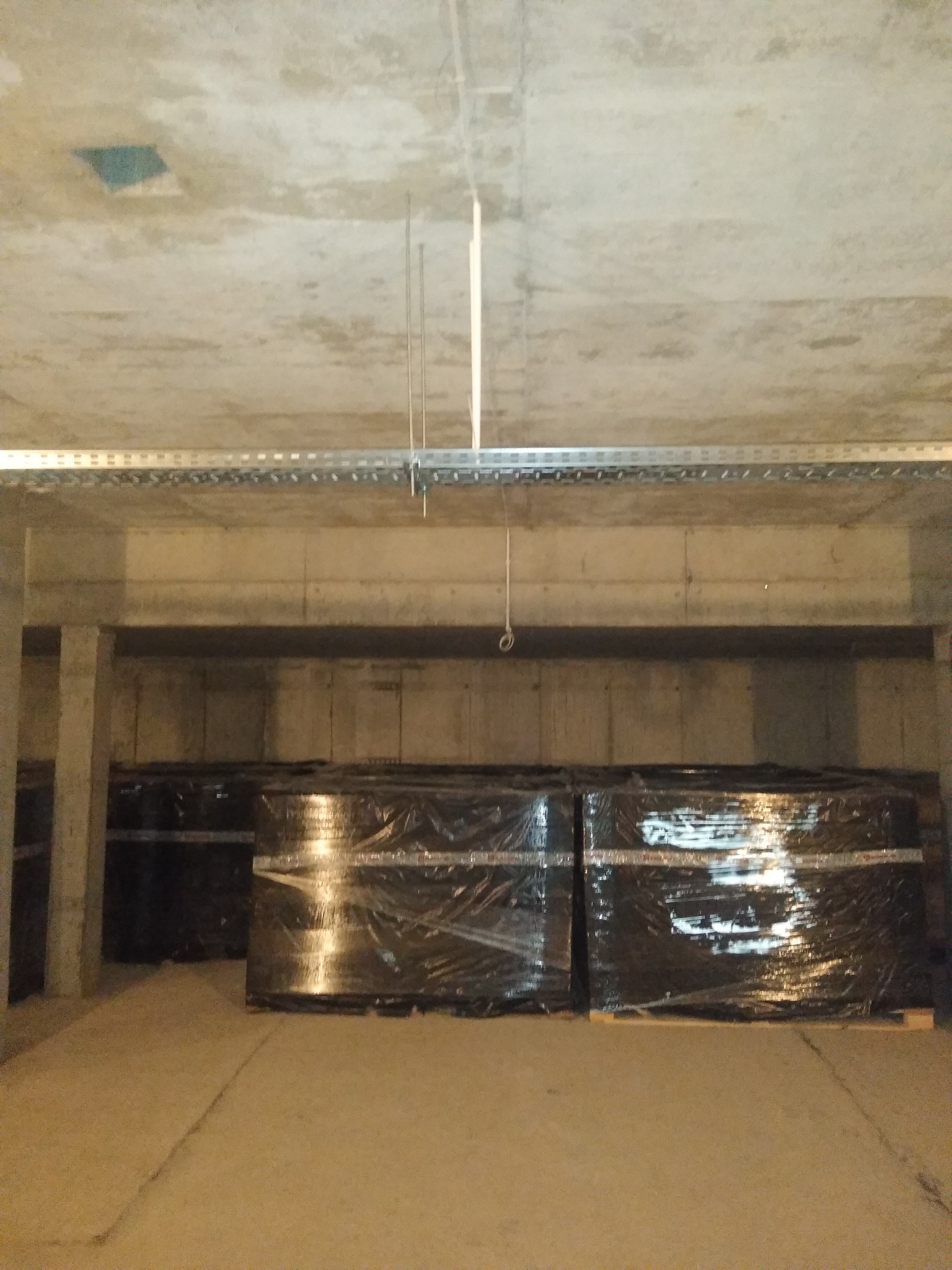 Sufit surowy z metalową szyną i zwisającymi przewodami, poniżej palety z materiałami budowlanymi w folii, w pomieszczeniu o betonowych ścianach.