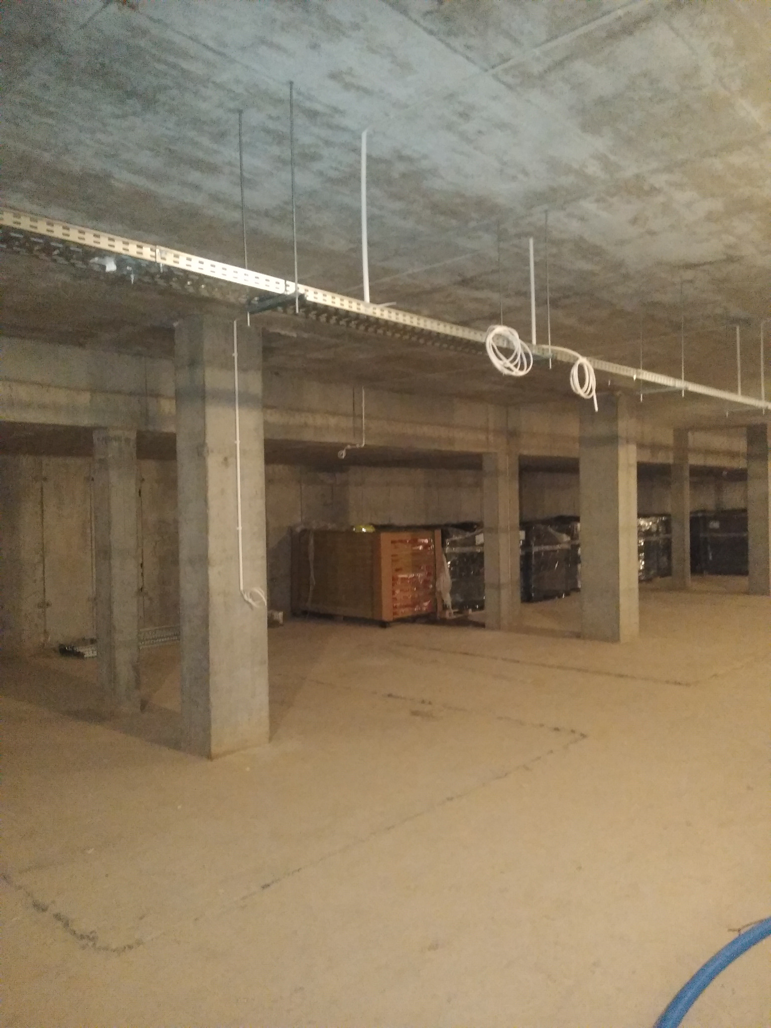 Betonowe słupy podtrzymują sufit z zamontowanymi korytkami kablowymi i przewodami. W tle widoczne materiały budowlane składowane w przestrzeni.