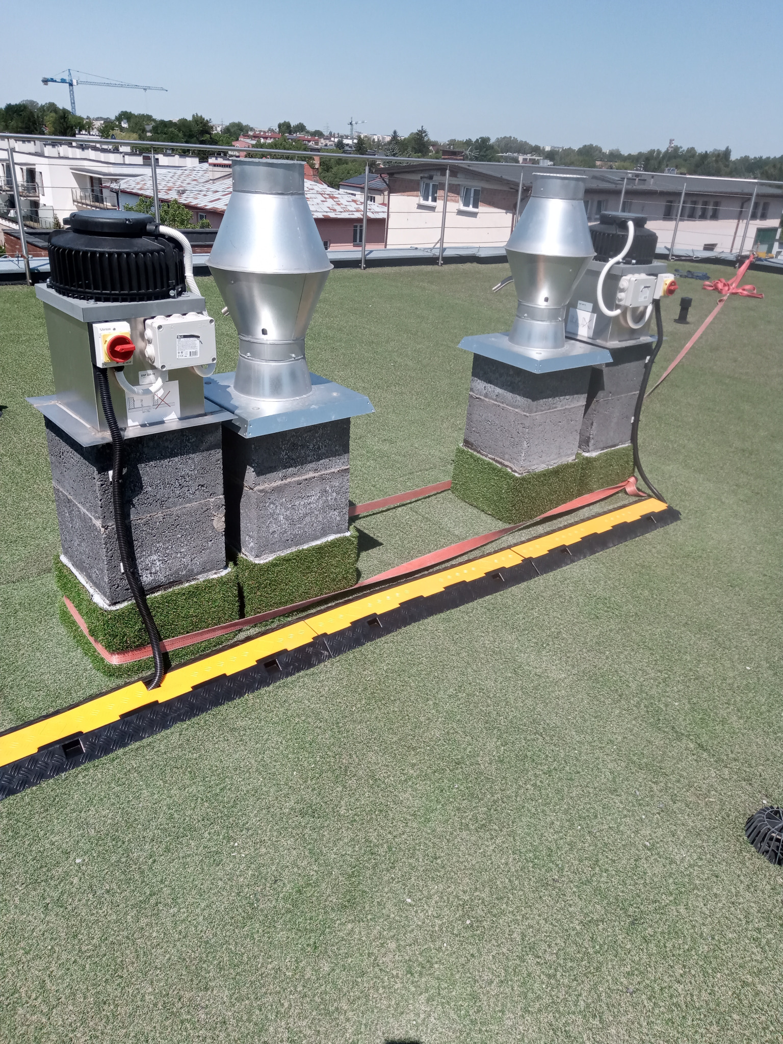 Dwa kominy wentylacyjne z silnikami na dachu pokrytym sztuczną trawą, zabezpieczone żółto-czarnymi osłonami kablowymi i czerwonymi taśmami. W tle budynki i błękitne niebo.