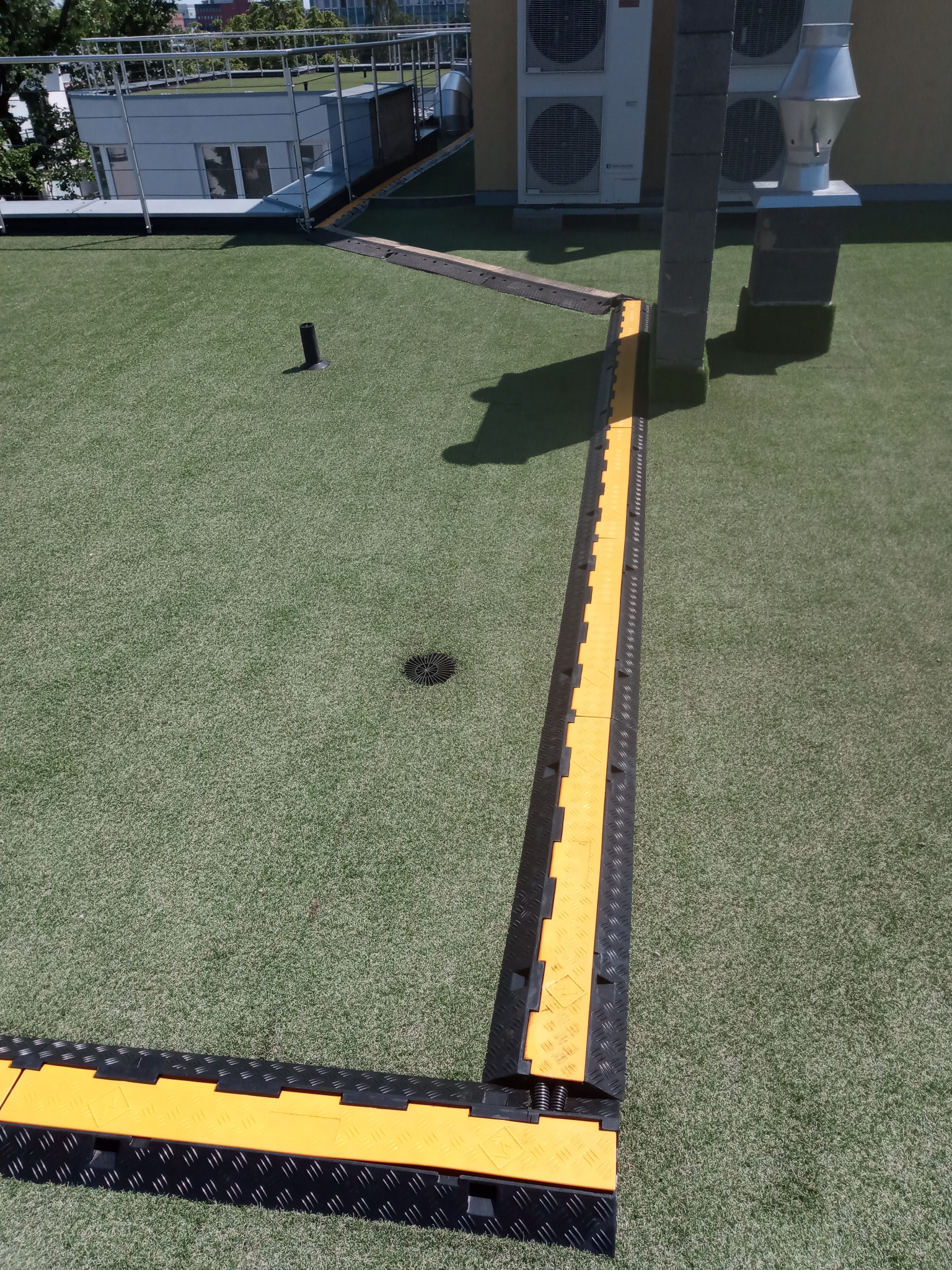 Żółto-czarne korytka kablowe ułożone na sztucznej trawie na dachu budynku, zabezpieczające przewody. Widoczne klimatyzatory i elementy konstrukcyjne dachu.