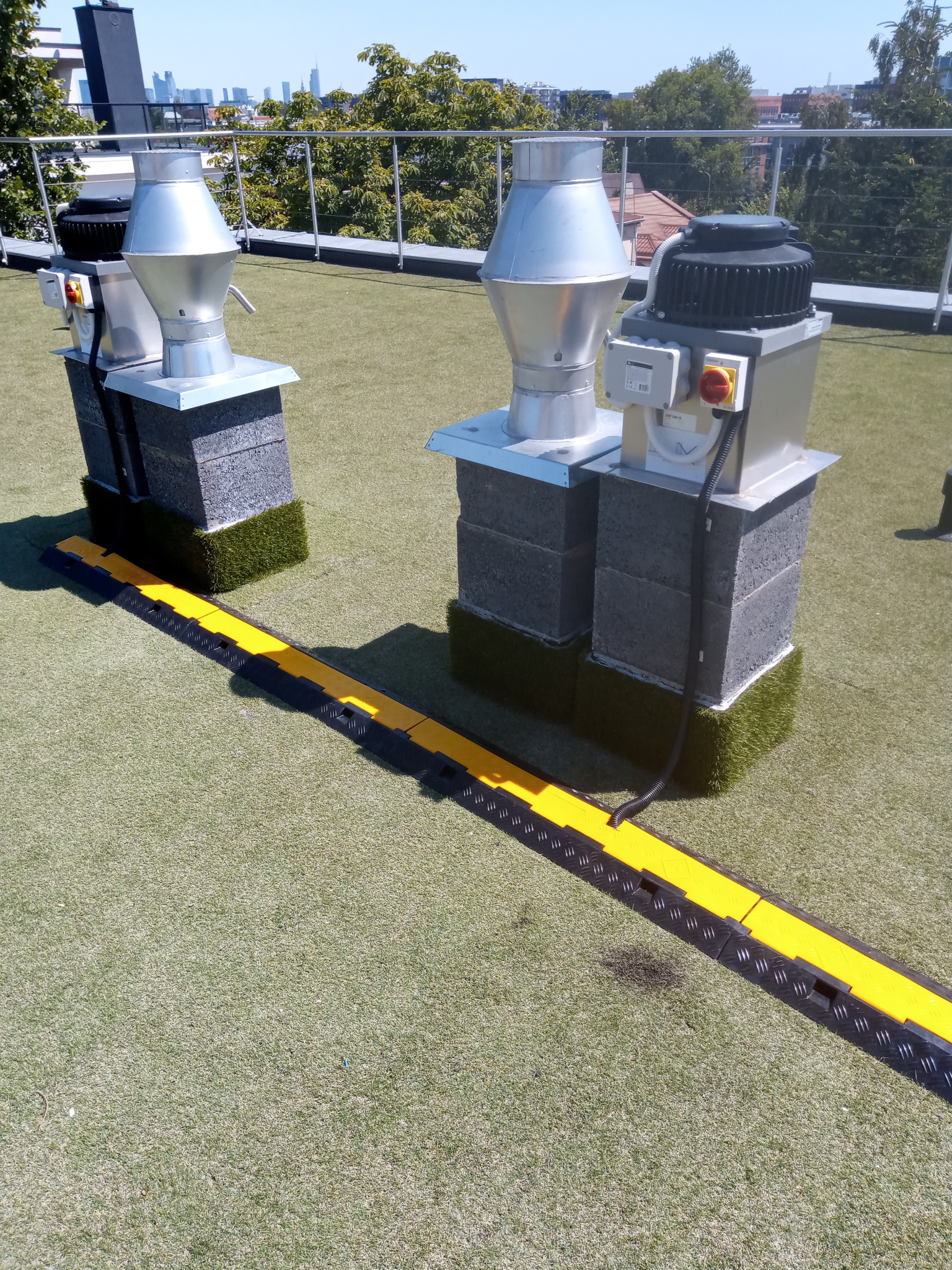 Dwa wentylatory przemysłowe na dachu pokrytym sztuczną trawą, zabezpieczone żółto-czarną osłoną kablową. Widok z góry, w tle budynki miejskie.