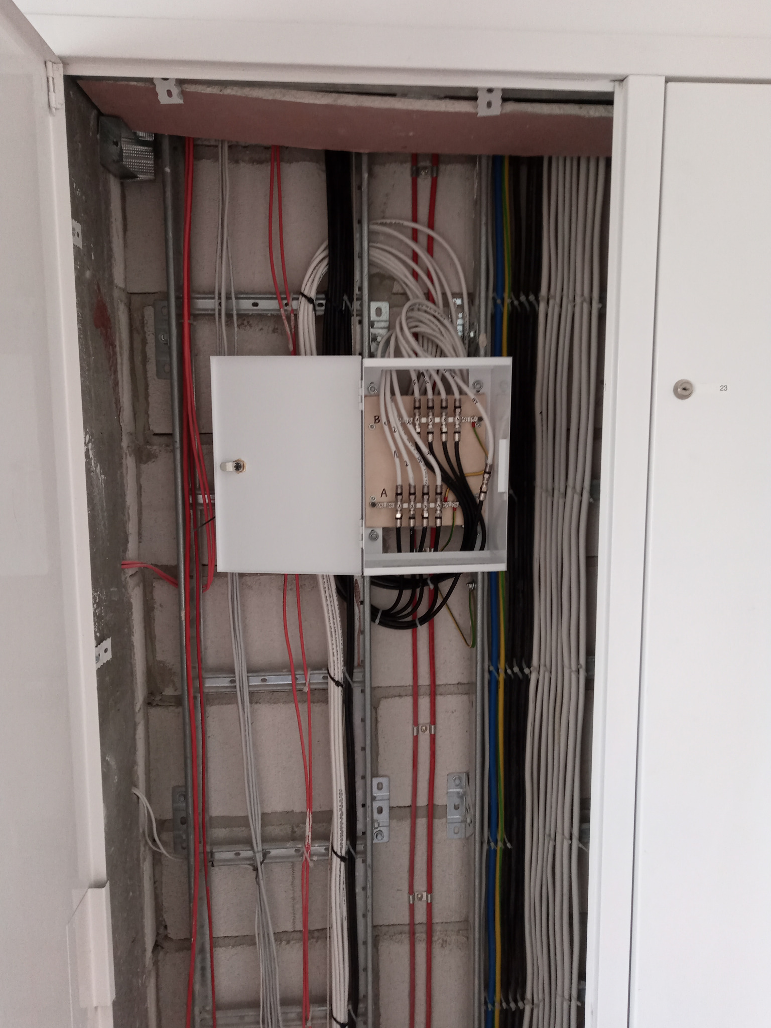 Otwarta skrzynka z kablami antenowymi wewnątrz szafy instalacyjnej. Widoczne kable w różnych kolorach, ułożone wzdłuż ściany z betonu komórkowego.