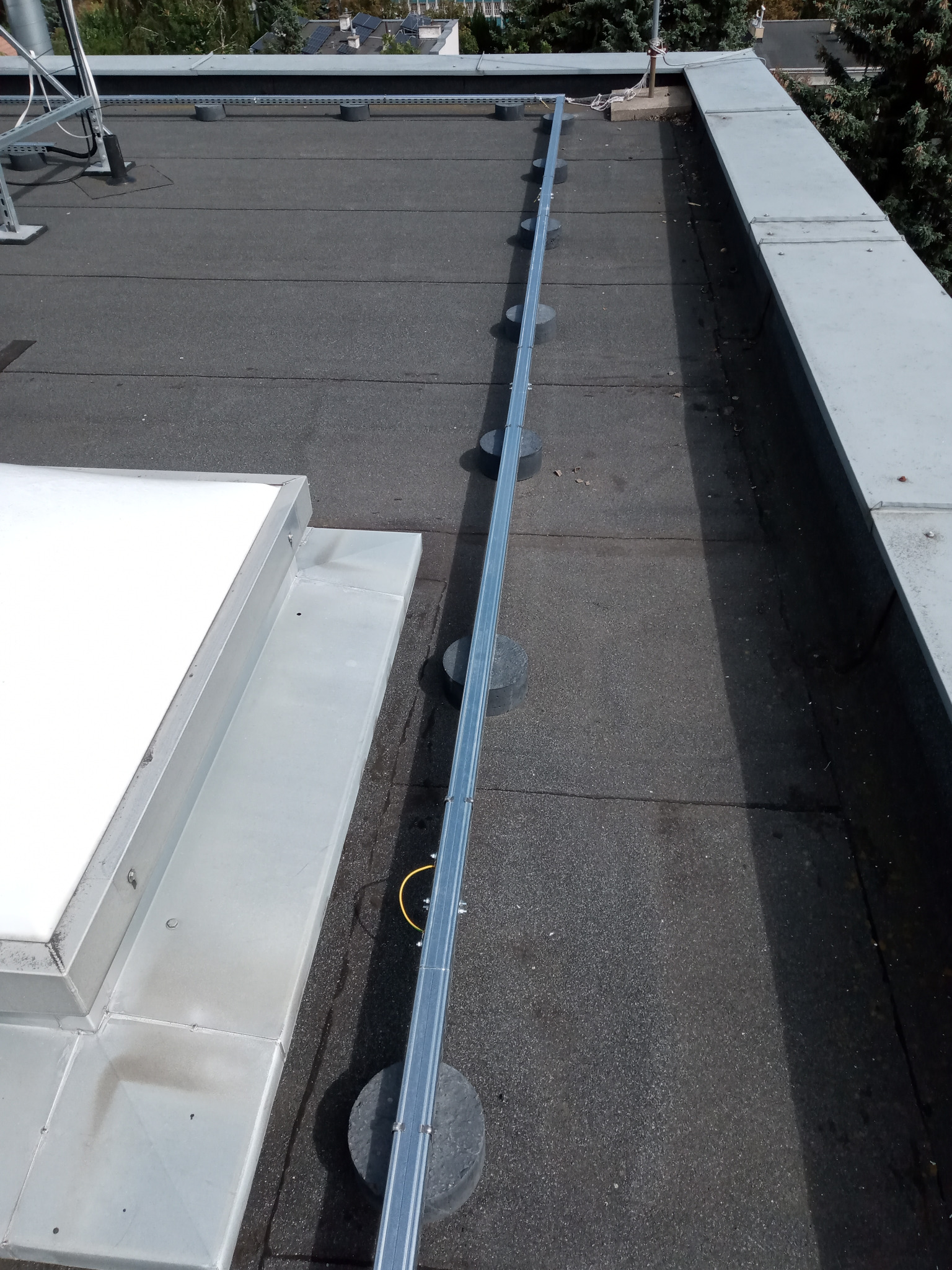 Szara listwa instalacyjna na czarnym dachu budynku, zamocowana na betonowych podstawach. Widok z góry, oświetlenie słoneczne.