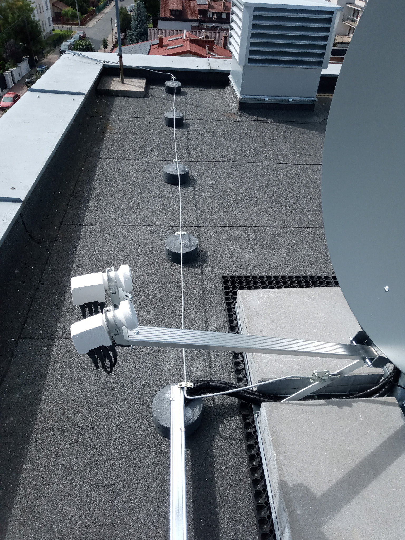 Anteny satelitarne na dachu pokrytym papą, zabezpieczone odgromowo. Widoczne uchwyty, przewody i elementy mocujące. Ujęcie z góry, w tle zabudowa miejska.