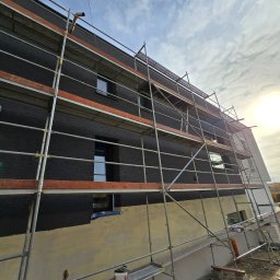 Przemek Najlepszy - Elewacja domu w trakcie prac wykończeniowych z czarną cegłą klinkierową, widoczne rusztowania, fragment ściany z nałożonym tynkiem podkładowym, błękitne niebo z chmurami w tle.