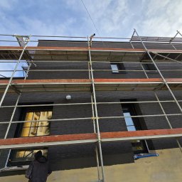 Przemek Najlepszy - Pracownik w czarnej kurtce na rusztowaniu maluje elewację budynku na ciemny kolor, widoczne okna i niebo z chmurami.