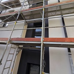 Przemek Najlepszy - Elewacja budynku w trakcie prac wykończeniowych, widoczne rusztowanie, fragmenty tynku o różnej fakturze i kolorystyce, okna z czarnymi ramami.